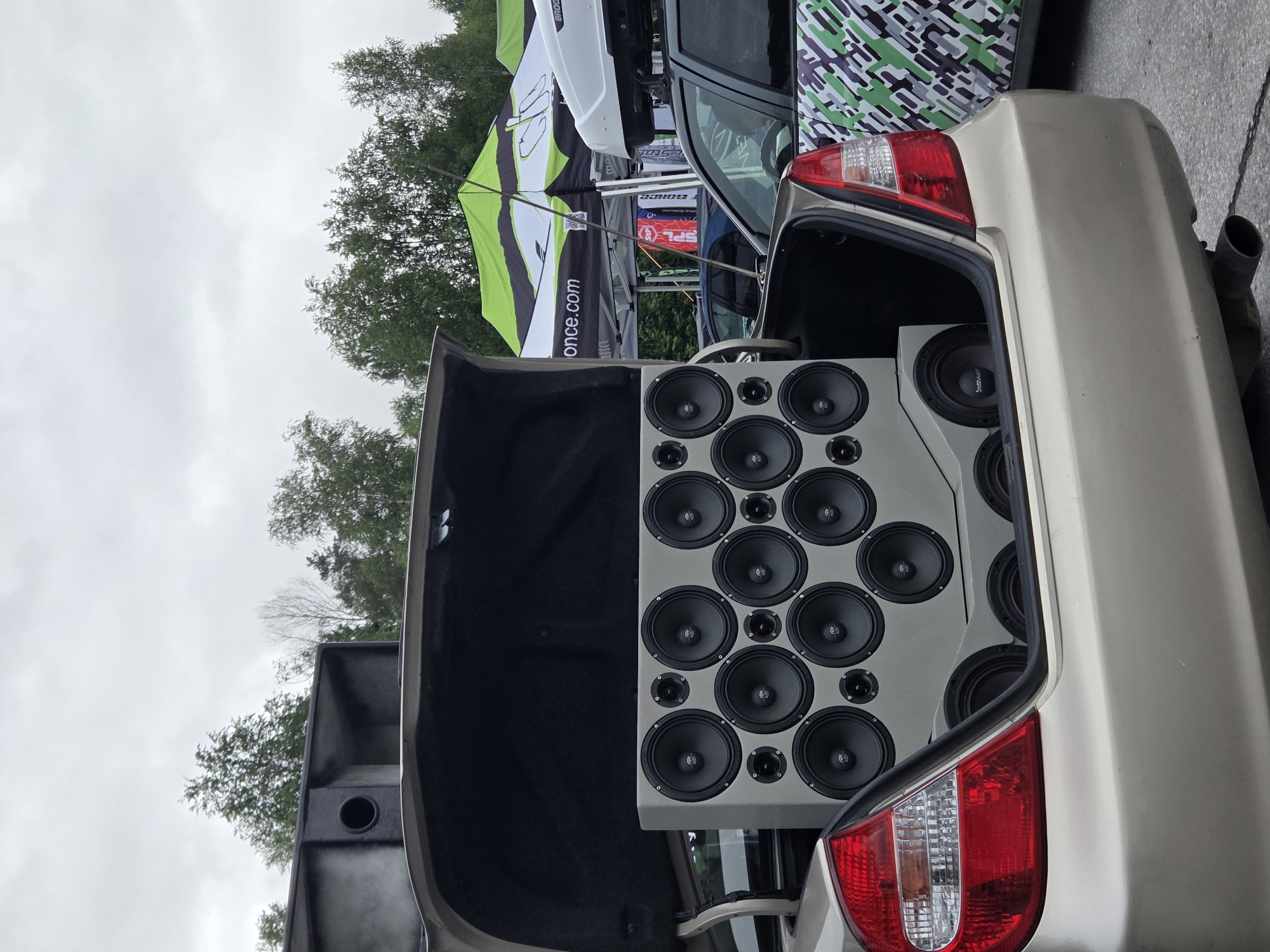 An outdoor scene in Perm, Russia, depicts the rear section of a gold-colored sedan with its trunk open, showcasing an extensively customized car audio system. The vehicle, possibly a Toyota Camry, is oriented with its rear towards the viewer. Inside the open trunk, a large, custom-fabricated light grey enclosure is filled with numerous circular speaker drivers. The visible portion of the enclosure features an array of 12 smaller speakers interspersed with 6 larger speakers, all bearing a stylized 'S' logo on their dust caps. Below this main panel, additional speakers are visible, including one marked "Skar Audio" and another with "FaSWAT" lettering. Above the speaker enclosure, within the trunk lid's structure, a black, horn-loaded audio component or port is integrated.
The environment is an overcast day, with a grey sky visible above a line of green deciduous trees in the background. Behind and to the right of the sedan, green and white striped event tents are erected, bearing partial text such as "once.com" and "Bondo." A dark-colored vehicle with a roof cargo box (partially labeled "BROOX") and a distinctive green, grey, and black geometric pattern wrap on its side is parked immediately to the right of the gold sedan. The ground is paved asphalt. No individuals are visible in the frame.