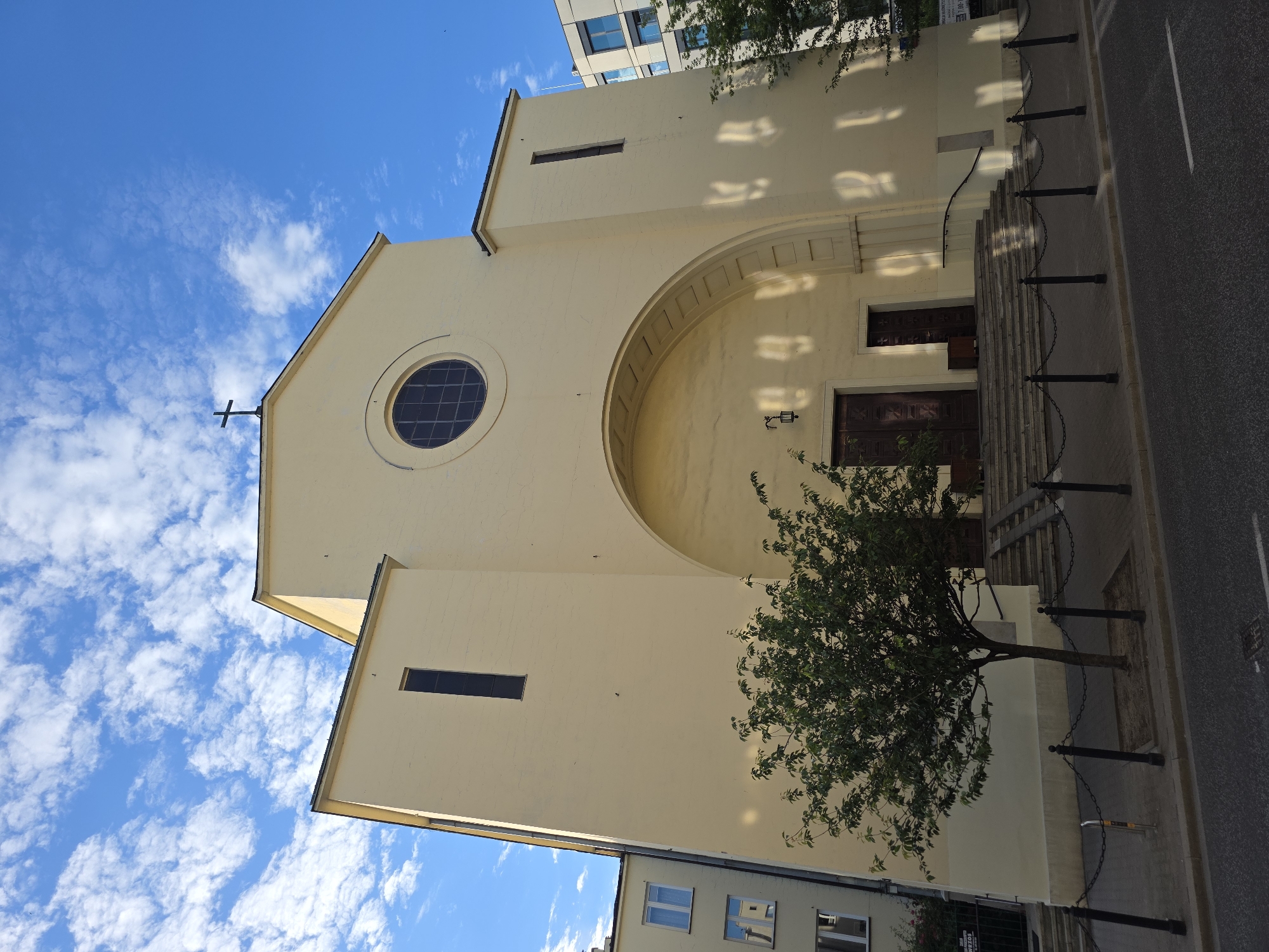 The image shows a pale yellow church with a dark cross on its roof, located in the city of Warsaw, Poland. The church has a large arched entrance with a set of stairs leading up to two wooden doors. A circular window is visible above the arch. To the left of the church, a modern apartment building with white windows is visible. A tree with green leaves stands in front of the church, separating it from the sidewalk and a street. The sky is blue with scattered white clouds. The overall scene is a clear, daytime shot of a prominent religious building in an urban setting.