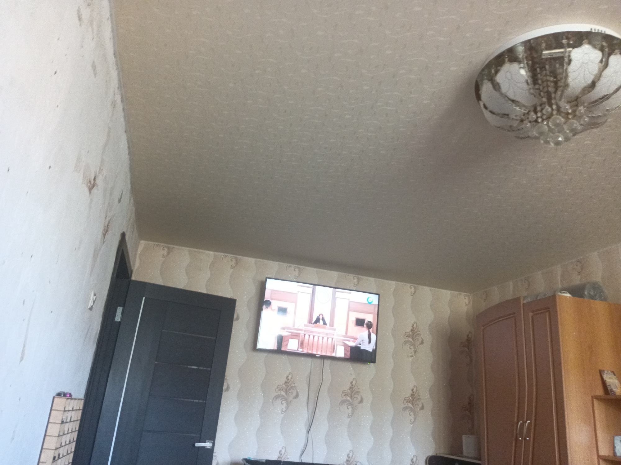 The image shows a residential interior, likely a living room in Kuznetsk, Russia. The scene features a dark grey door on the left side of the frame, with a thin white vertical stripe. To the right of the door, a television is mounted on a wall covered in patterned wallpaper. The TV displays a scene from what appears to be a courtroom drama, with people seated at desks and a central figure at a podium.
In the upper right portion of the image, a ceiling light fixture with a classic, ornate design is visible, featuring clear crystal accents and a white lampshade. Further to the right, a tall wooden cabinet or wardrobe stands against the wall, with two doors that have curved detailing. The wallpaper in this area also has a repeating pattern, featuring floral or scroll motifs.
The walls appear to be a light color, with some areas showing signs of wear or imperfections, particularly on the left side of the frame where the plaster seems to be exposed in places. The overall lighting suggests it is daytime, though the specific time of day is not evident from the image. There are no visible signs of weather conditions. The room appears to be a private residence. No specific emotions are conveyed by the scene. There is no visible text in the image.