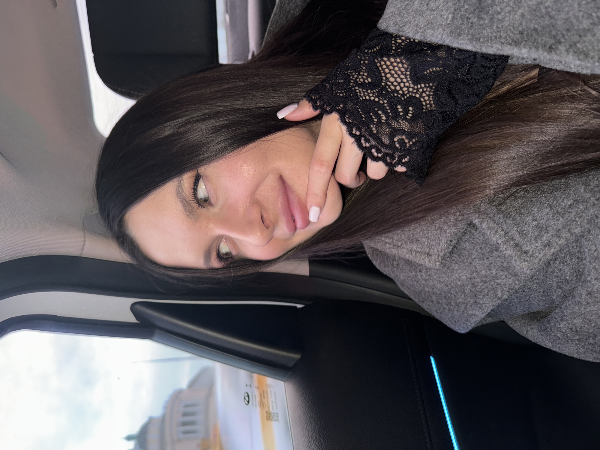 A brunette woman is sitting in a car in Moscow, Russia. She is wearing a gray coat and black lace sleeves. Her face is turned to the side, and she is smiling. The reflection of a building is visible outside of the window.