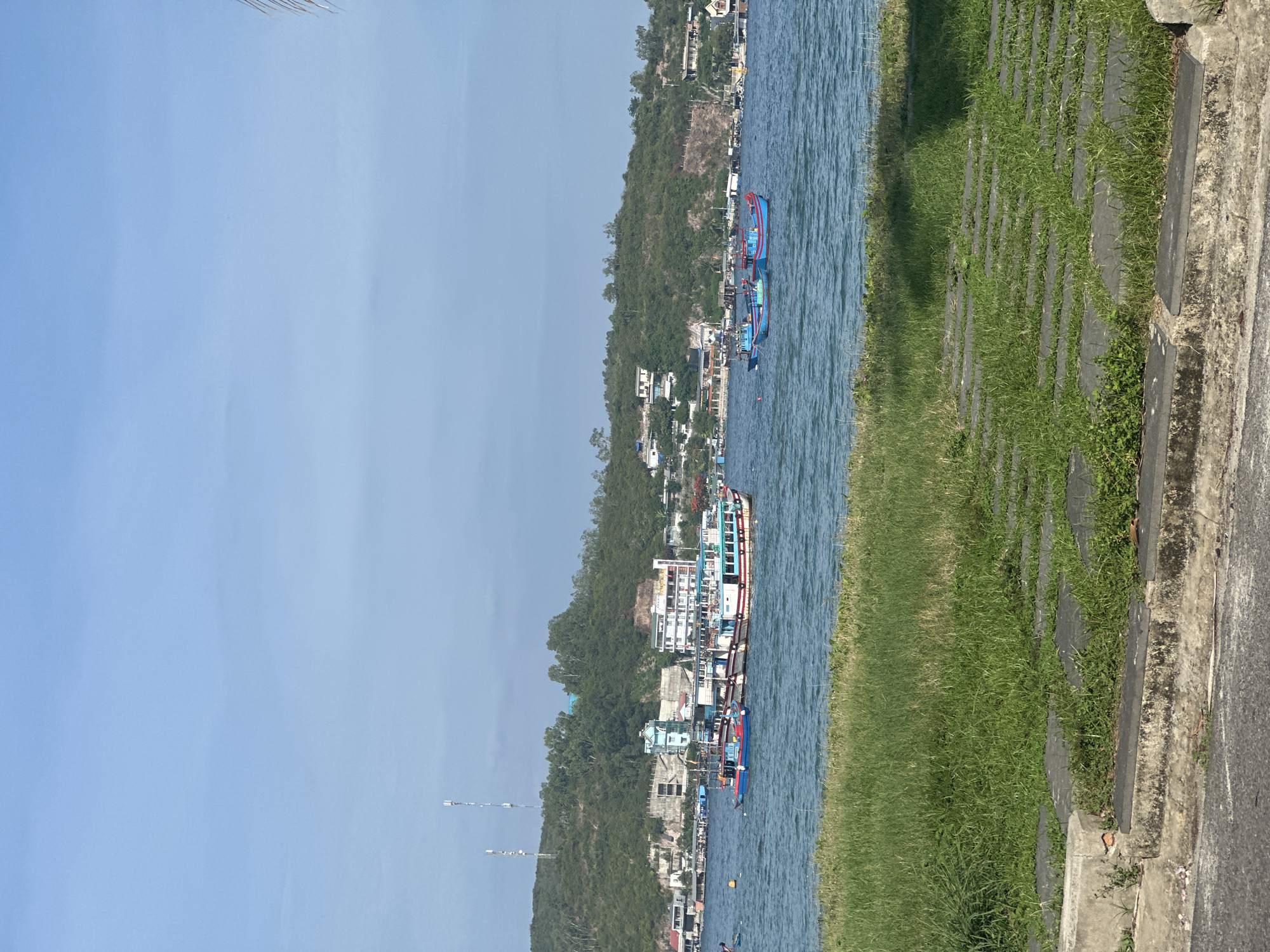 This image depicts a coastal scene in Nam Nha Trang Ward, Vietnam. The foreground features a paved pathway with grass growing between the stones, bordering a stretch of green grass. Beyond the grass, a body of water with moderate waves is visible. In the distance, several boats are present in the water. Some of these boats are larger, with multi-colored hulls and white cabins, suggesting they might be fishing vessels or passenger boats. Smaller, brightly colored boats are also scattered across the water. The background reveals a lush, green hillside dotted with buildings and some prominent communication towers. The sky is a clear, pale blue with a few wispy clouds.