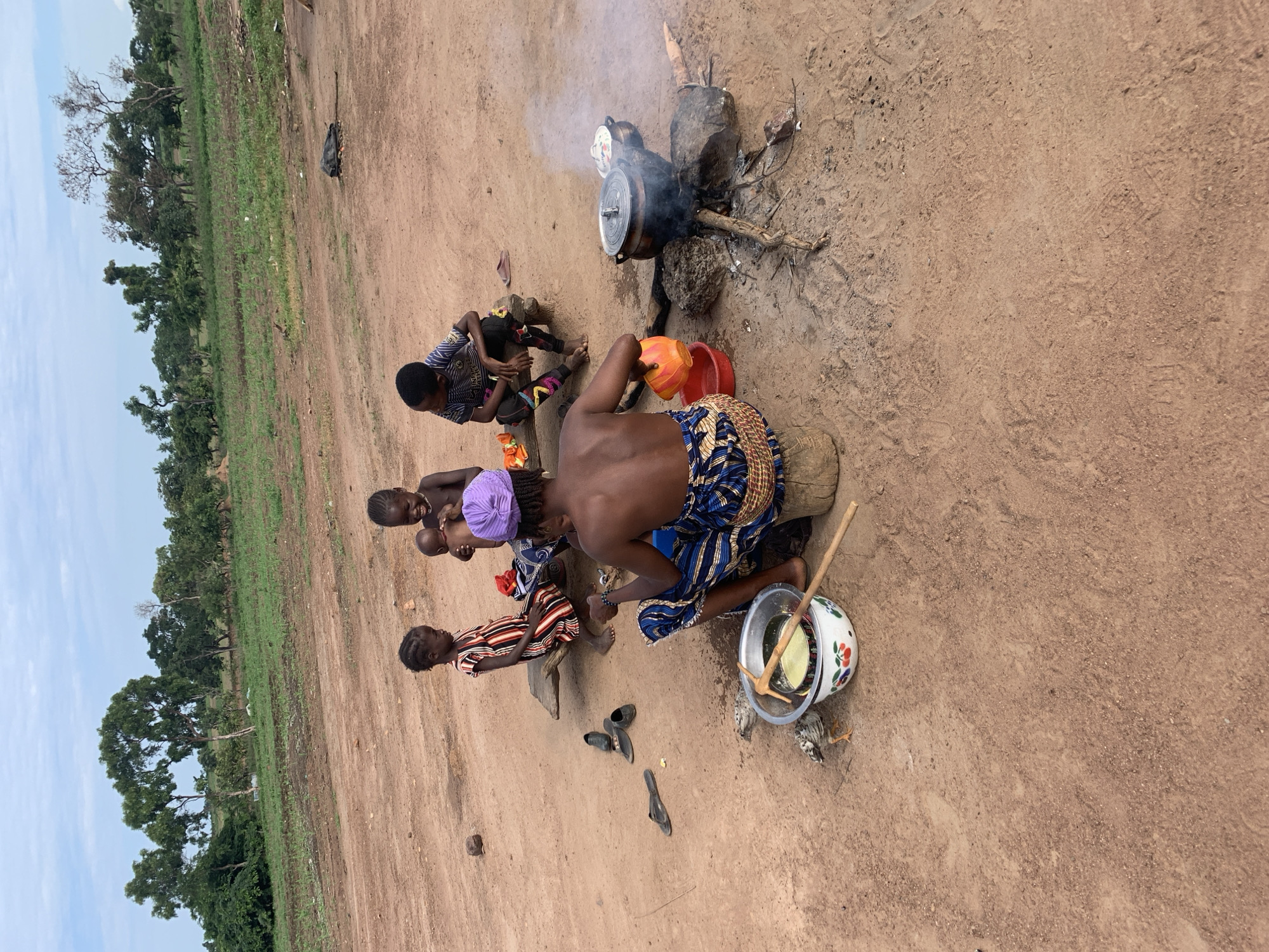 In Nandu, Nigeria, a group of individuals gathers around an outdoor cooking fire. A woman, wearing a blue and white patterned skirt and a purple headwrap, is seated on a log, tending to a cooking pot over the fire. She holds an orange bowl in her right hand, and a red bowl is near her left. To her left, a metal bowl with a cherry pattern contains a yellow liquid and a wooden utensil. Two young children are seated on a wooden plank to the left of the woman. One child, dressed in a striped dress, is looking down, while the other, holding a baby, is looking towards the camera and smiling. A third child, wearing a striped shirt and pants, sits further down the plank, looking to the right. Several pairs of shoes are scattered on the ground. In the background, a field with greenery and trees stretches towards the horizon under a cloudy sky. A black object, possibly a rolled-up mat or bag, lies on the ground in the middle distance. The ground is predominantly red dirt.