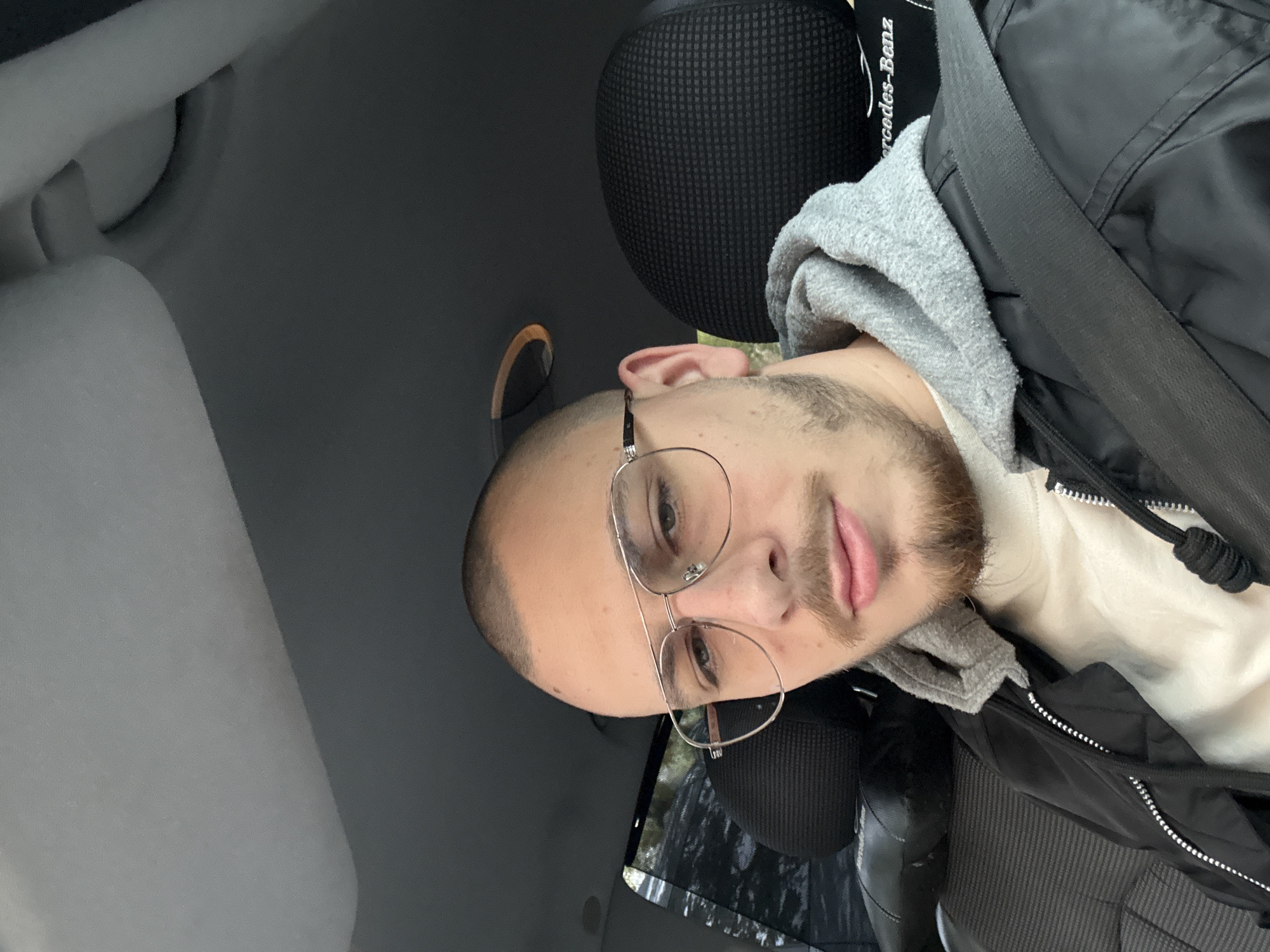 A tilted, eye-level photograph captures a young man seated inside a car, likely in Stara Zagora, Bulgaria. The man, positioned towards the right side of the frame, has a shaved head, a light beard, and a mustache. He is wearing round, thin-framed glasses and has a neutral to slightly smirking expression while looking towards the camera.
He is dressed in a light-colored top, a grey hooded sweatshirt, and a black jacket. A black bag or backpack with a strap is visible over his right shoulder, resting against the car's headrest and his jacket. The interior of the car is visible, featuring grey and black tones. The seat headrest, made of a textured black material, clearly displays the white text "Mercedes-Benz," indicating the car's make. The lighting suggests it is daytime, casting soft illumination on the scene.