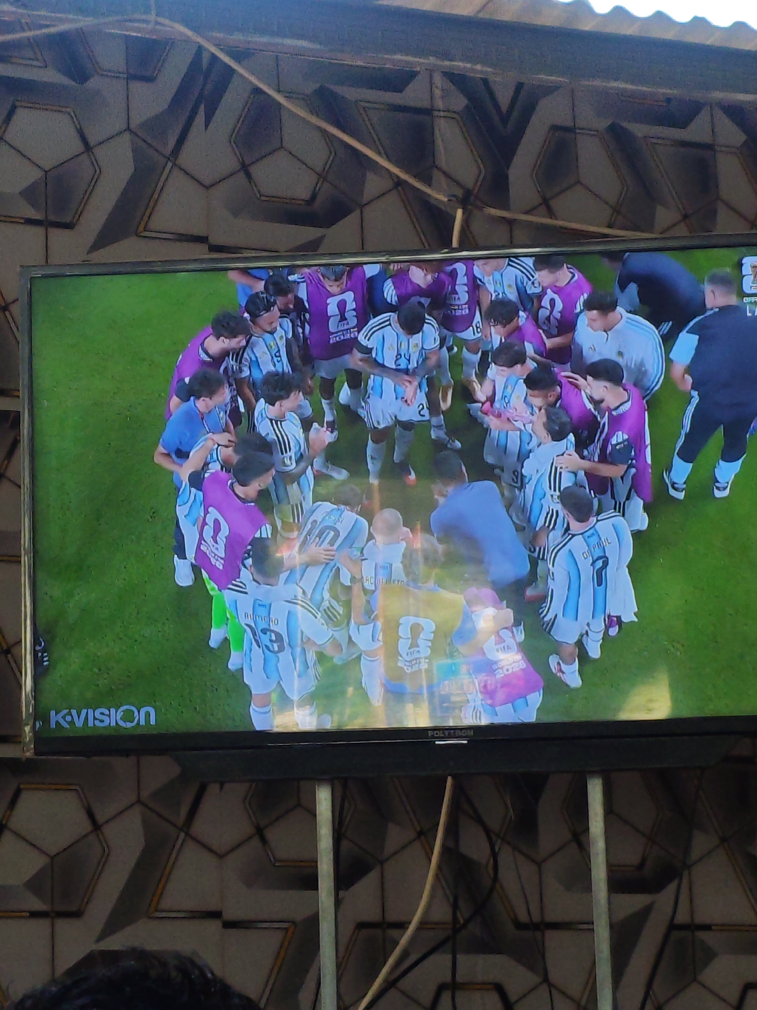 A television screen displays a group of young men in white and blue striped soccer uniforms, some wearing purple vests over their jerseys, huddled together on a green field. Their arms are around each other, suggesting celebration or camaraderie. The television is mounted on a wall with a geometric pattern in shades of gray, white, and black. The bottom edge of the television shows branding: "KVISION" on the left and "POLYTRON" on the right. The scene appears to be viewed from a slightly elevated angle. This image was captured in Parangsembilan, Indonesia.