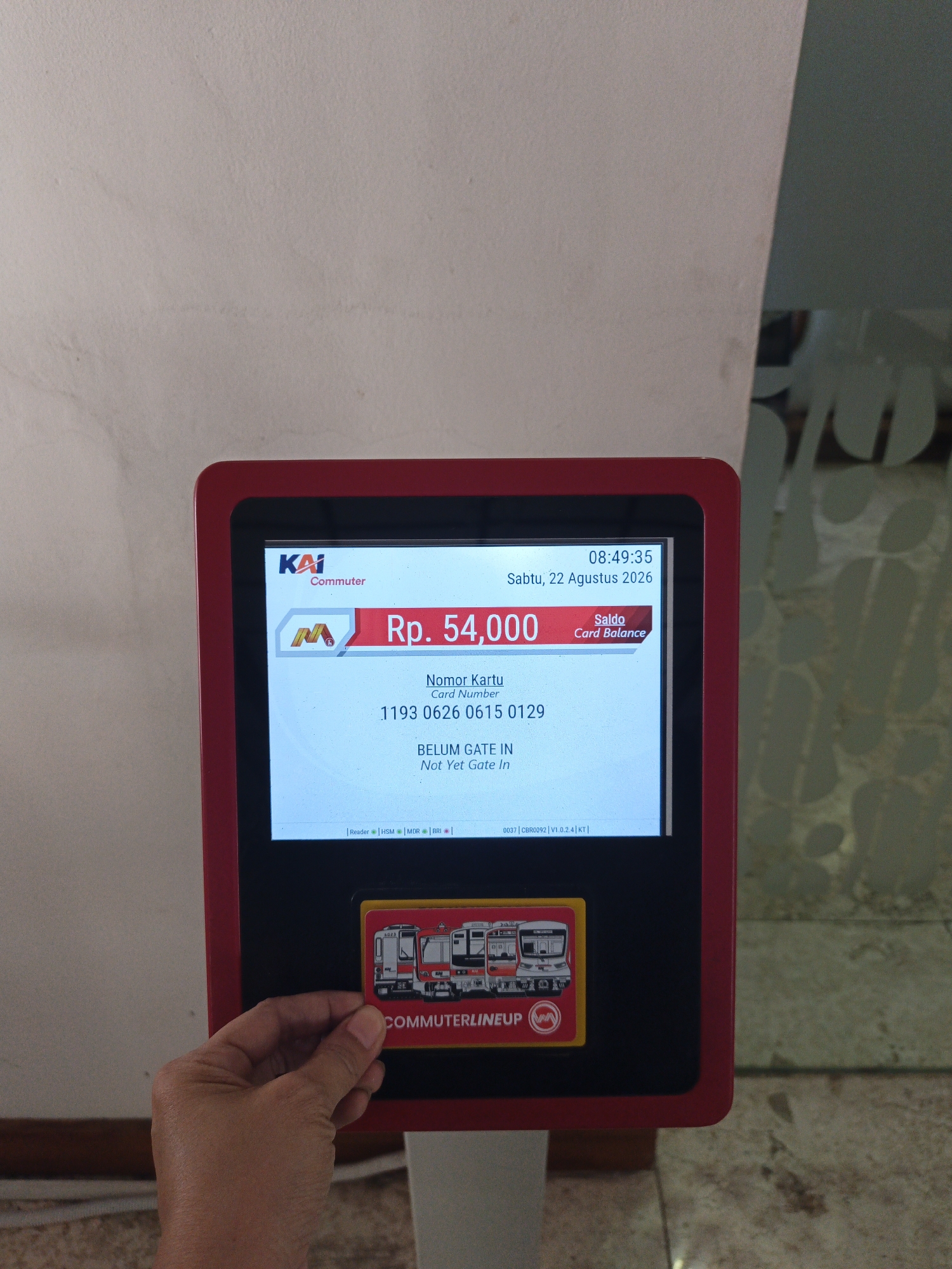 The image shows a close-up of a red-framed electronic payment terminal with a screen displaying information. A hand is holding a commuter pass with an image of trains and the text "COMMUTERLINEUP" towards the screen. The screen shows the KAI Commuter logo, a balance of "Rp. 54,000", a card number "1193 0626 0615 0129", and a message "BELUM GATE IN | Not Yet Gate In". The time displayed is "08:49:35" and the date is "Sabtu, 22 Agustus 2026" (Saturday, August 22, 2026). Below the main display, there are smaller text labels indicating "Reader", "HSM", "MDR", and "BRI". The terminal is positioned indoors, with a blurred background showing a textured wall and a glass partition with a faded pattern. The image depicts a scene at a transportation hub, likely a train station, where commuters use these terminals to check their balance and pass through gates. While the image provides details about the transaction, it does not offer specific information to pinpoint the exact location within Klaten, Indonesia, other than implying a public transportation setting.