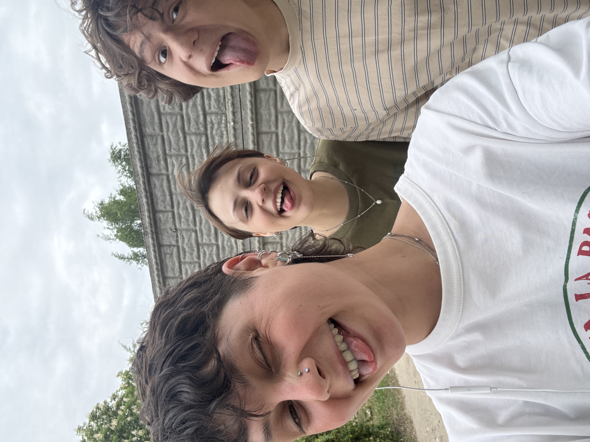 Three people are posing for a selfie. The photo is taken from a low angle, looking up at the subjects. The person in the foreground is wearing a white t-shirt and has dark curly hair. They have a nose piercing and multiple ear piercings. They are sticking their tongue out. The person in the middle is wearing a green shirt and a silver necklace. They are also sticking their tongue out. The person in the background is wearing a tan and white striped shirt and has curly brown hair. They are sticking their tongue out.
In the background, there is a concrete wall. There are some trees and greenery visible. The sky is overcast. It is impossible to verify if the location is Uryupinsk, Russia.