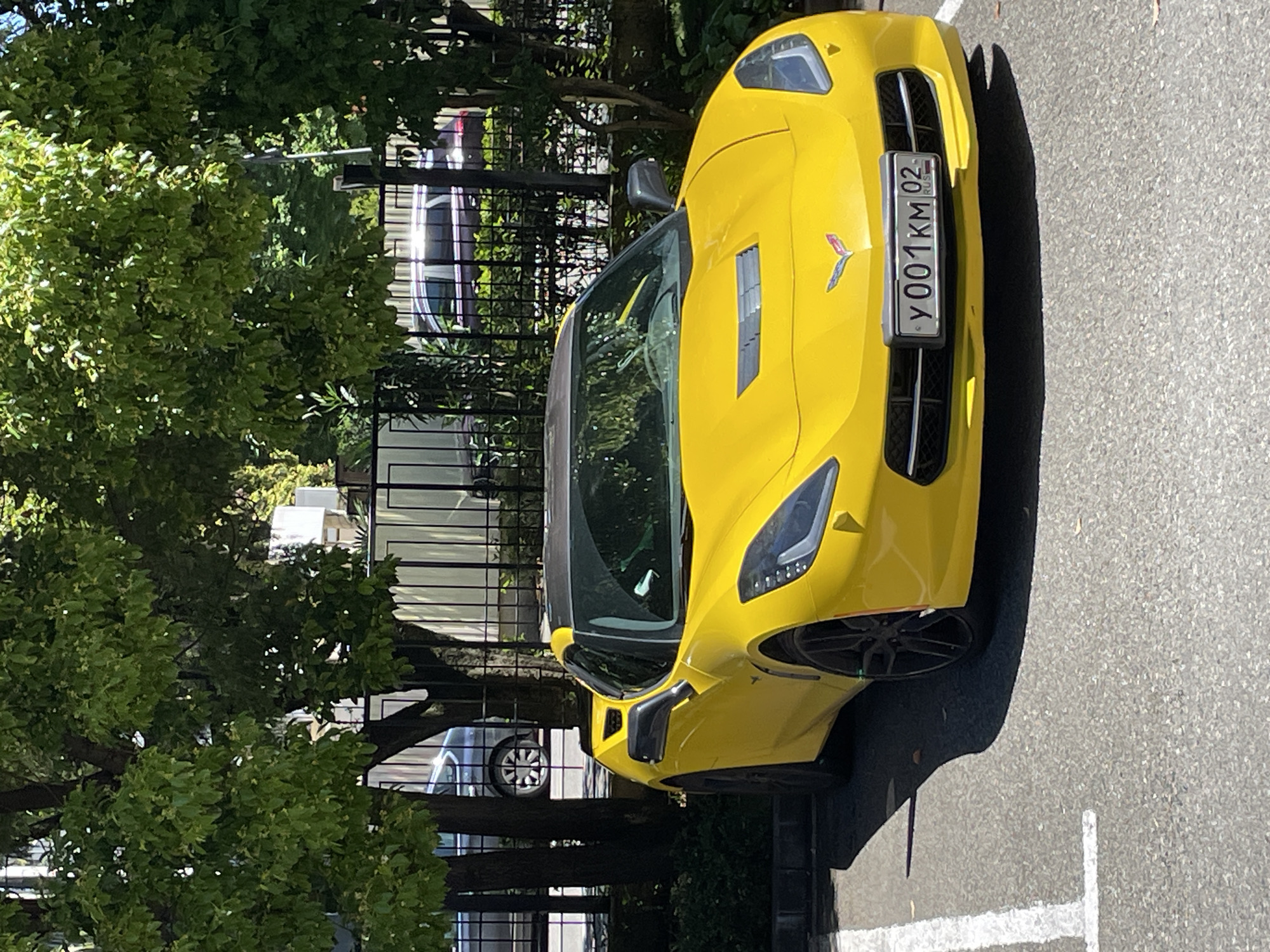 A bright yellow Chevrolet Corvette, likely a C7 generation model, is prominently parked outdoors on an asphalt surface in Sochi, Russia. The sports car faces slightly towards the viewer, showcasing its aggressive front fascia, distinctive headlights, and a small Corvette emblem on the hood. A black front license plate, "Y001KM 02 RUS", is clearly visible in the center of the grille, indicating its Russian origin with region code "02".
The scene is bathed in bright sunlight, casting strong, well-defined shadows to the right side of the car, suggesting a clear and sunny daytime. The car's vivid yellow paint reflects the light, highlighting its sculpted lines and air vents on the hood and side panels.
In the background, dense, lush green trees and foliage create a natural, verdant backdrop. A black metal fence runs horizontally behind the vehicle, separating the parking area from the treeline. Through the gaps in the foliage and fence, parts of other parked cars and some building structures are subtly visible. The overall setting appears to be a parking lot or street-side parking area, consistent with an urban or resort environment like Sochi. No people are visible, and the car is stationary.