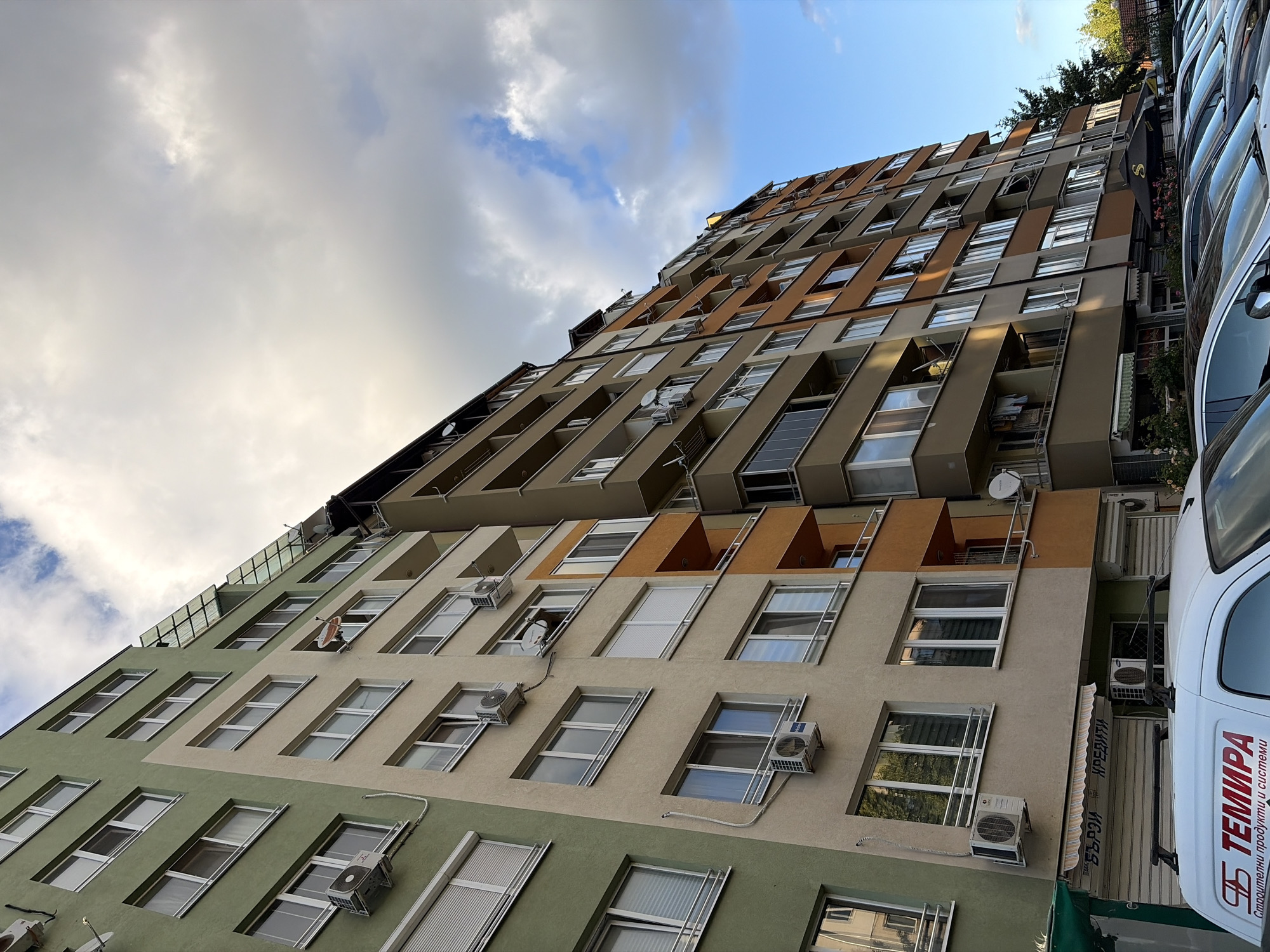 The image shows a tall, multi-story apartment building with a mix of green, beige, and orange facade colors. Several windows and air conditioning units are visible on the building's exterior. The sky above is partly cloudy with patches of blue sky. In the foreground, the roof of a white van with red lettering is visible, along with a row of parked cars. Some trees and foliage are also present on the right side of the image. The scene appears to be an urban or suburban environment.