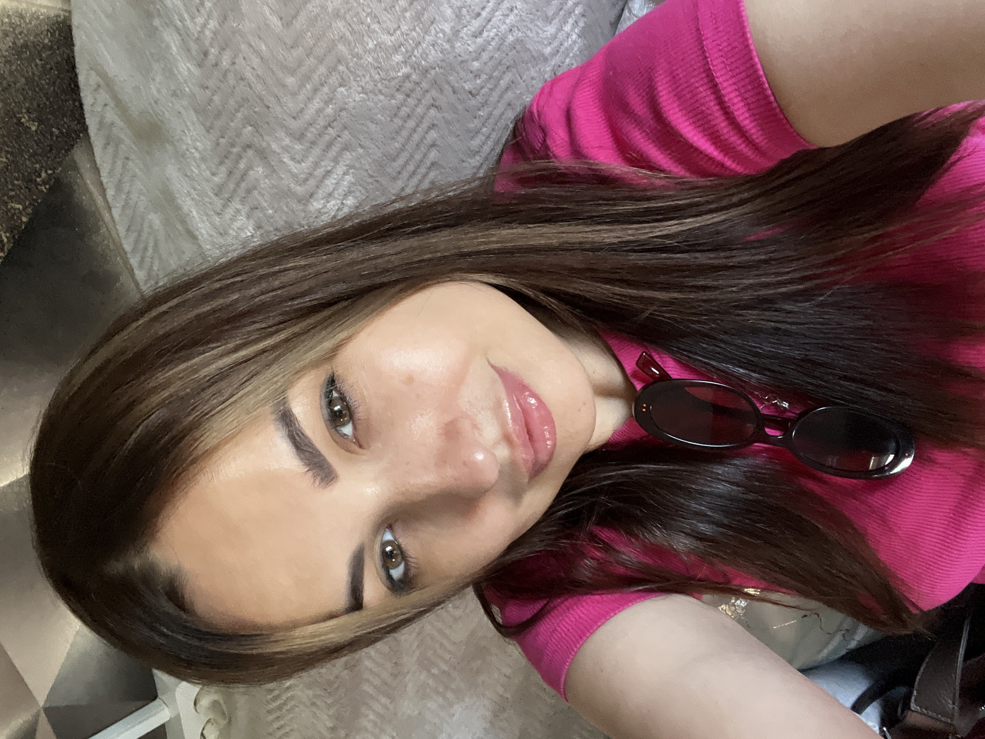 A woman is taking a selfie indoors, likely on a sofa or bed, given the textured fabric visible in the background. She has long, dark brown hair with lighter brown highlights, and she is wearing a bright pink ribbed short-sleeved shirt. A pair of dark sunglasses with red accents is hanging from the neckline of her shirt. Her makeup includes lip gloss and eyeliner. The lighting suggests an indoor environment, possibly during the day.