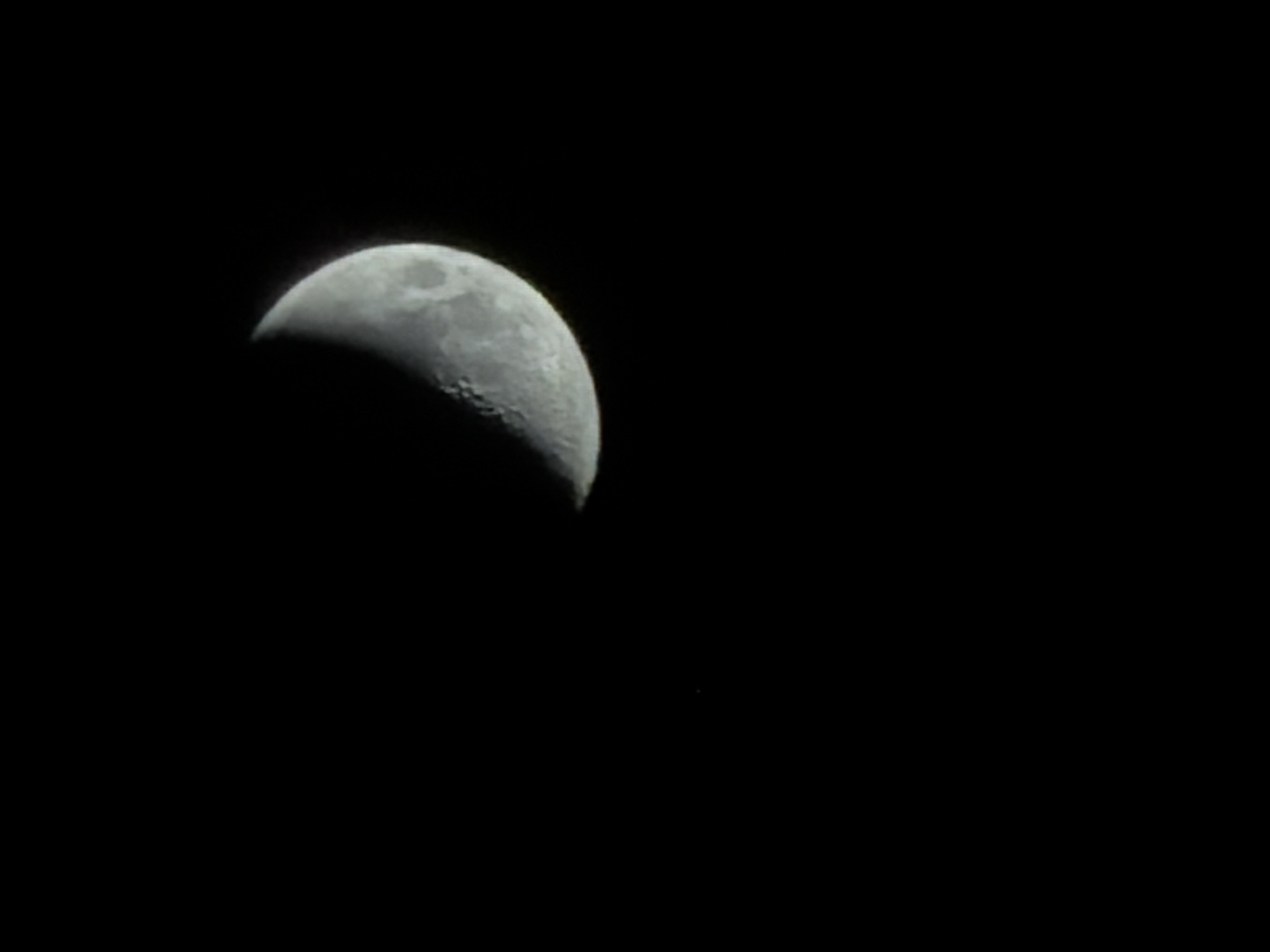 The image displays a clear, close-up photograph of a crescent moon against a completely black night sky. The moon, illuminated on its right side, appears to be in a waxing phase, likely a first quarter or early waxing gibbous. Its surface details are visible, including numerous craters and darker plains (maria), particularly evident along the terminator line separating the lit and shadowed portions. The texture of the lunar surface is well-defined. The moon occupies the left-central area of the frame, with vast expanse of dark sky to its right. A very faint, tiny white speck, possibly a distant star or image artifact, is barely visible in the lower right portion of the dark background. The scene suggests a clear, cloudless night, allowing for unobstructed astronomical observation. There are no other discernible celestial bodies, landscape features, or visible text. The photograph captures a tranquil view of the moon from Izhevsk, Russia.