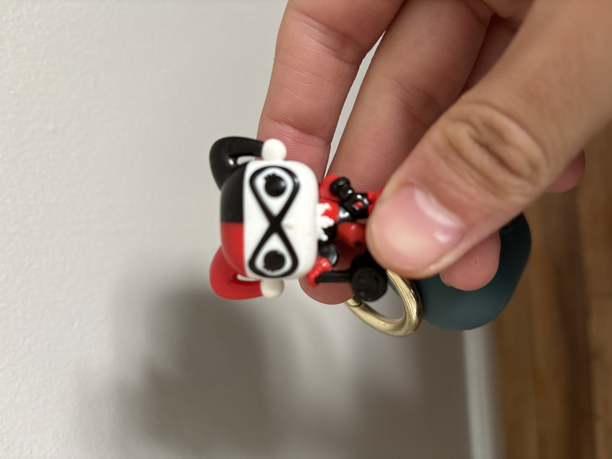 A person is holding a cartoon keychain. The keychain features a character with a split black-and-white design on its head and body. The white side has two black eye-like shapes, and the red side has a smaller figure riding on its back. The keychain is attached to a gold-colored ring and a dark green object. The photograph appears to be taken indoors against a blurred background. It is impossible to determine the location of this scene with the information provided.