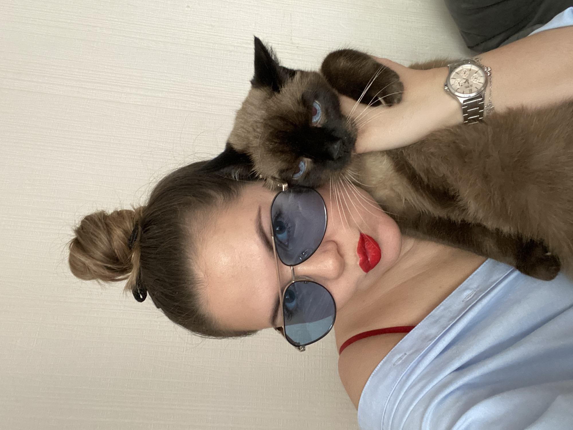 A woman with a cat is in a room. The woman has her hair in a bun and is wearing sunglasses and red lipstick. She is wearing a light blue shirt with a red string around her neck. The cat is a Siamese cat with blue eyes. The woman has a watch on her left wrist. The background is a plain, light-colored wall. The image is taken from a close-up perspective. The location in the city of Izhevsk, Russia is unknown.