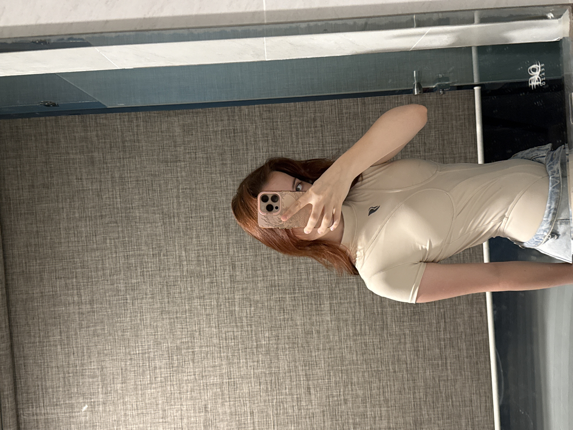The image shows a reflection of a person taking a selfie in a bathroom. The person has reddish-brown hair and is wearing a tan-colored athletic shirt and denim shorts. The bathroom wall is covered in a textured gray fabric. A mirror extends above the wall, with what appears to be the edge of a shower enclosure visible to the right. The location in the city of Bac Nha Trang Ward, Vietnam is not discernible from the image.