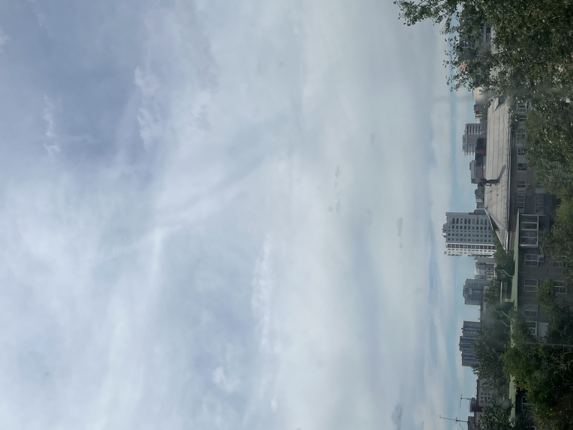 The image shows a cloudy sky over a city with apartment buildings and trees. The sky is filled with a mixture of white and gray clouds, with some lighter patches indicating breaks in the cloud cover. In the middle ground, several multi-story apartment buildings are visible, with varying architectural styles and heights. One prominent white building with many windows stands out. The foreground is dominated by lush green trees, with their leaves partially obscuring the lower floors of some buildings. The scene suggests a typical urban environment on a cloudy day.
This image was taken in Chelyabinsk, Russia.