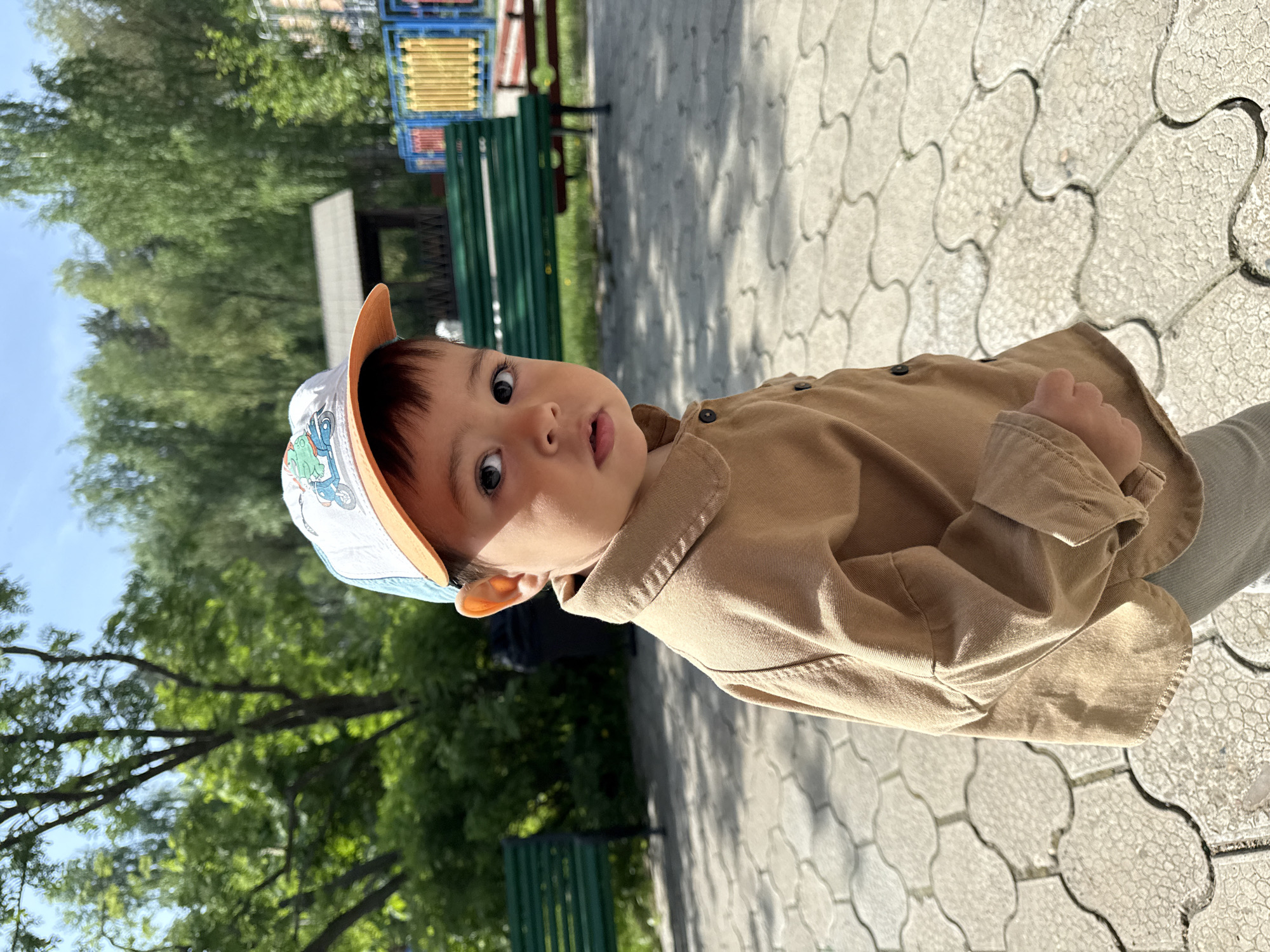 A young child, likely a toddler, stands on a paved surface in what appears to be a park or recreational area. The child is wearing a light brown button-down shirt and a cap with a light-colored front and a light orange brim. The paving stones are light gray and patterned. In the background, there are green trees, a green park bench, and a playground structure with multiple colors, including yellow, blue, and red. The time of day is likely daytime with sunlight present, casting shadows on the paving. The exact location in Chaikovsky, Russia, cannot be precisely determined from this image alone; however, based on the common features of a playground or park, it is plausible that this is a public park area.