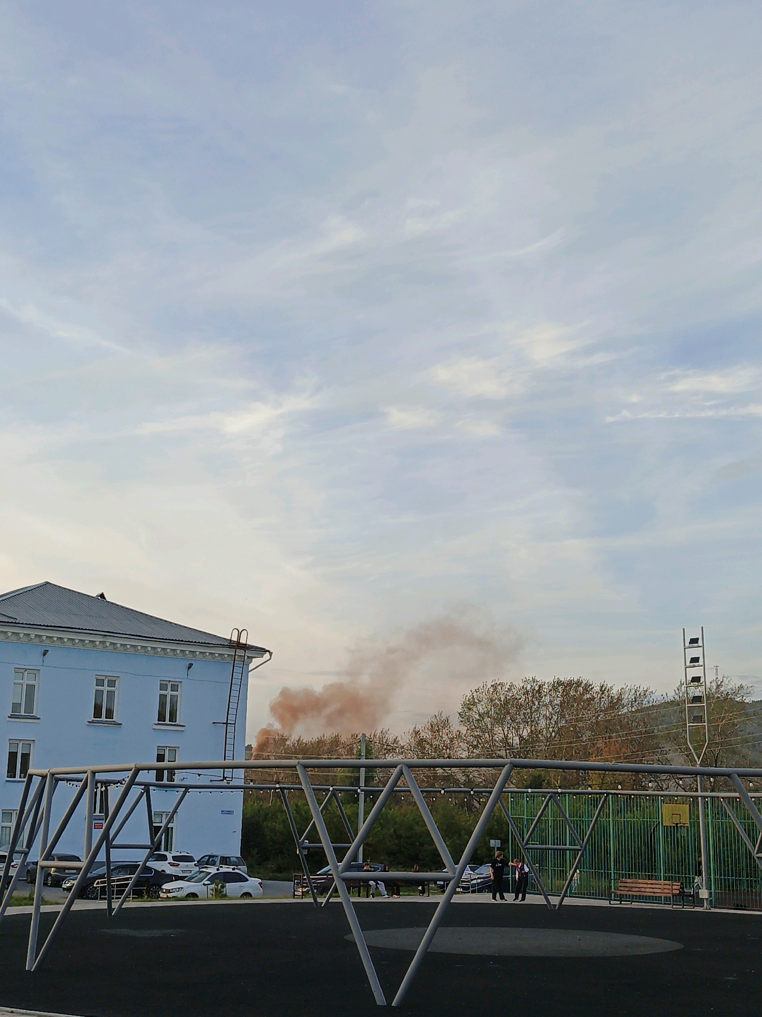 In Zlatoust, Russia, a light blue, multi-story building with a dark roof and white trim stands on the left side of the frame. A metal fire escape ladder is attached to the side of the building. In front of the building, a playground area with a black rubber surface features a metal swing set. Several cars are parked in front of the building. Beyond the playground, a line of trees with autumn foliage separates the area from a grassy hill in the background. A plume of reddish-brown smoke is visible rising from behind the trees. On the right side of the frame, a green metal fence encloses a basketball court with a yellow hoop. Two individuals, one in dark clothing and the other in a white shirt and dark pants, are standing near the playground. The sky above is filled with scattered, wispy clouds.
