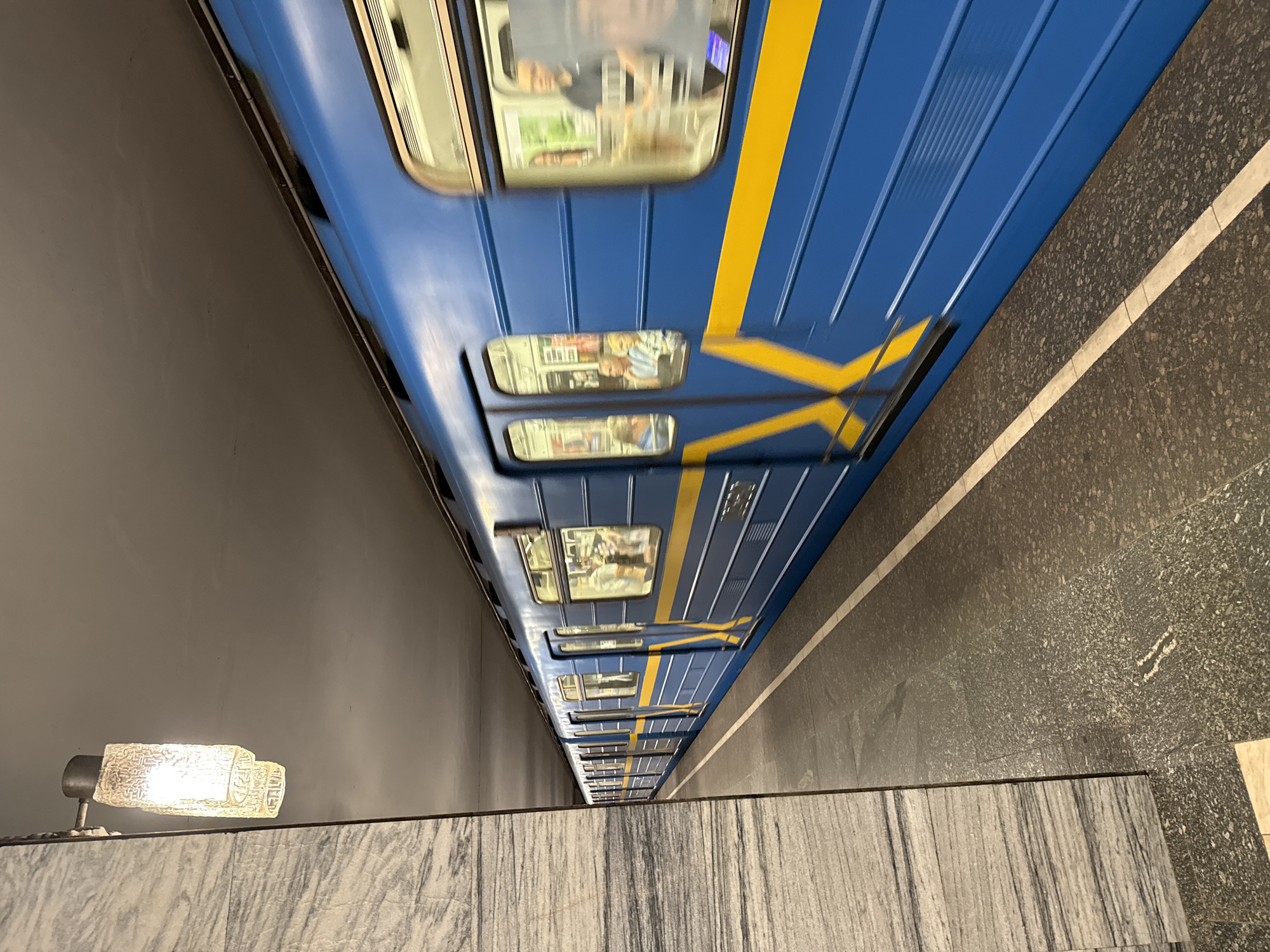 An eye-level, long shot captures a blue and yellow subway train moving from left to right in an underground station. The train is in motion, indicated by motion blur, and its windows reveal blurred figures of passengers. The station platform is dark gray with white lines demarcating the edge. To the left, a marble-clad pillar stands near a light fixture. The ceiling is dark and indistinct. The image's perspective is from the platform, looking down the length of the train as it departs.