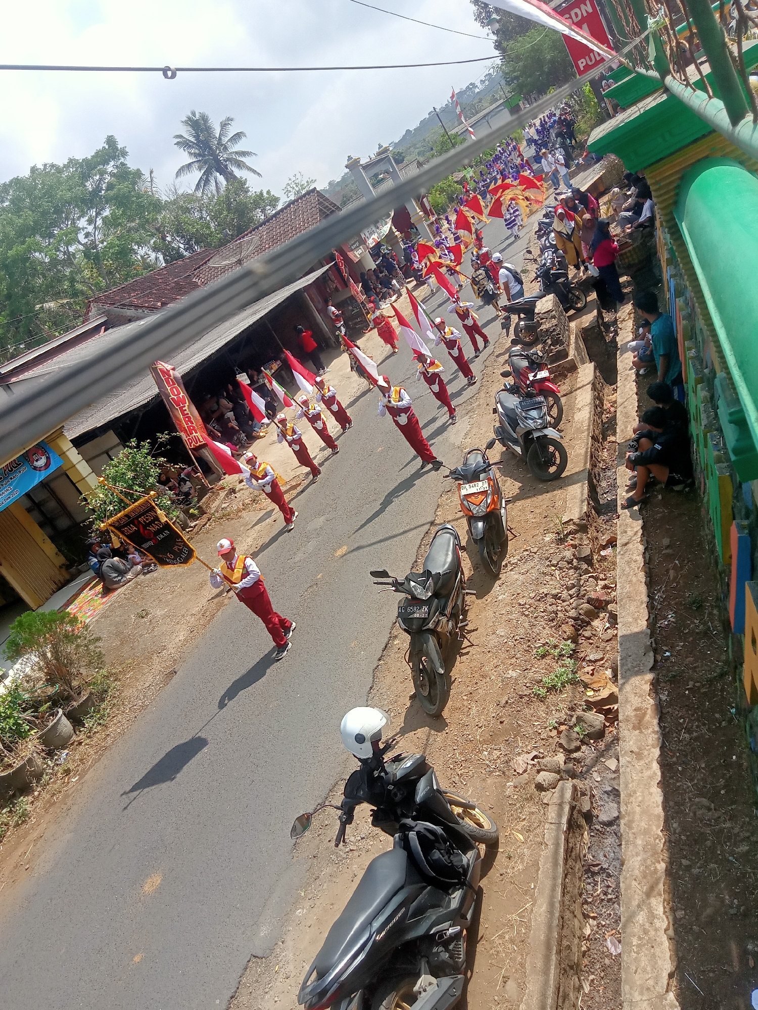 This image depicts a street scene in Pule, Indonesia, likely during a parade or procession. A group of individuals in matching red and white marching band uniforms are visible on the asphalt road, some holding flags or banners. They appear to be part of a school or local organization, as indicated by the banner reading "SDN 1 Pule" and "Jaya Preso Gnag". The parade route is lined with onlookers sitting and standing on the side of the road, some of whom are also holding Indonesian flags. Several motorcycles are parked along the roadside. The background shows buildings with tiled roofs, lush green trees, and a cloudy sky. The overall atmosphere suggests a community event or celebration.
