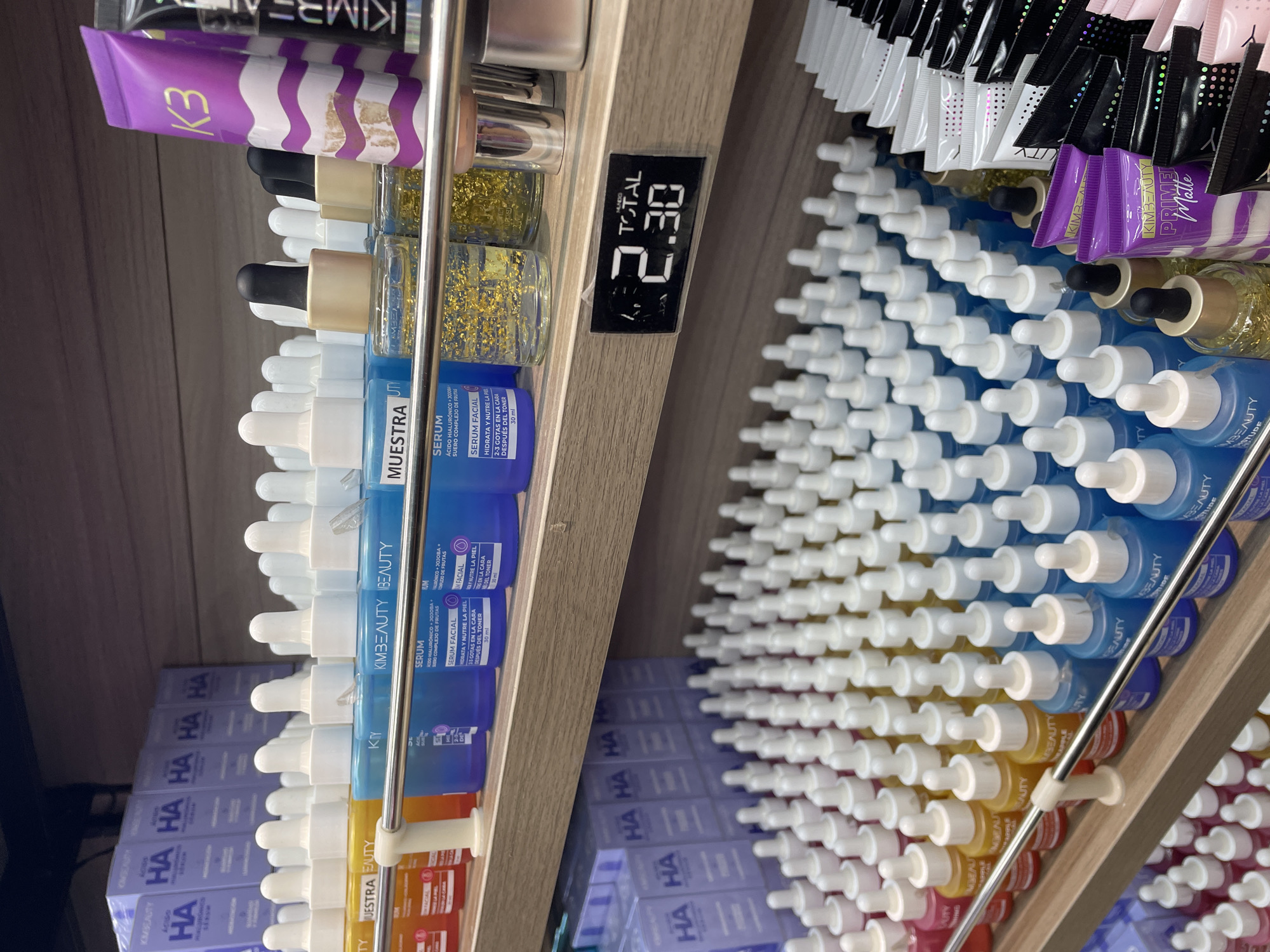 The image displays a retail shelf filled with various skincare products. The shelves are made of wood-like material and are organized with products in rows. In the foreground, there are rows of small glass bottles with droppers, containing liquids in different colors such as pink, yellow, and blue. Some of these bottles are labeled with "BEAUTY". Behind these, larger blue bottles labeled "SERUM" are visible. Further back and to the right, there are rows of purple tubes and black pouches, some with branding that includes "K3" and "KIMBEAUTY". A digital price tag showing "2.30" is attached to the shelf in the middle section. In the background, on a higher shelf, more purple boxes with "HA" branding are stacked. The overall environment appears to be a well-lit retail store or pharmacy section dedicated to beauty products. There are no people or animals visible in the image.