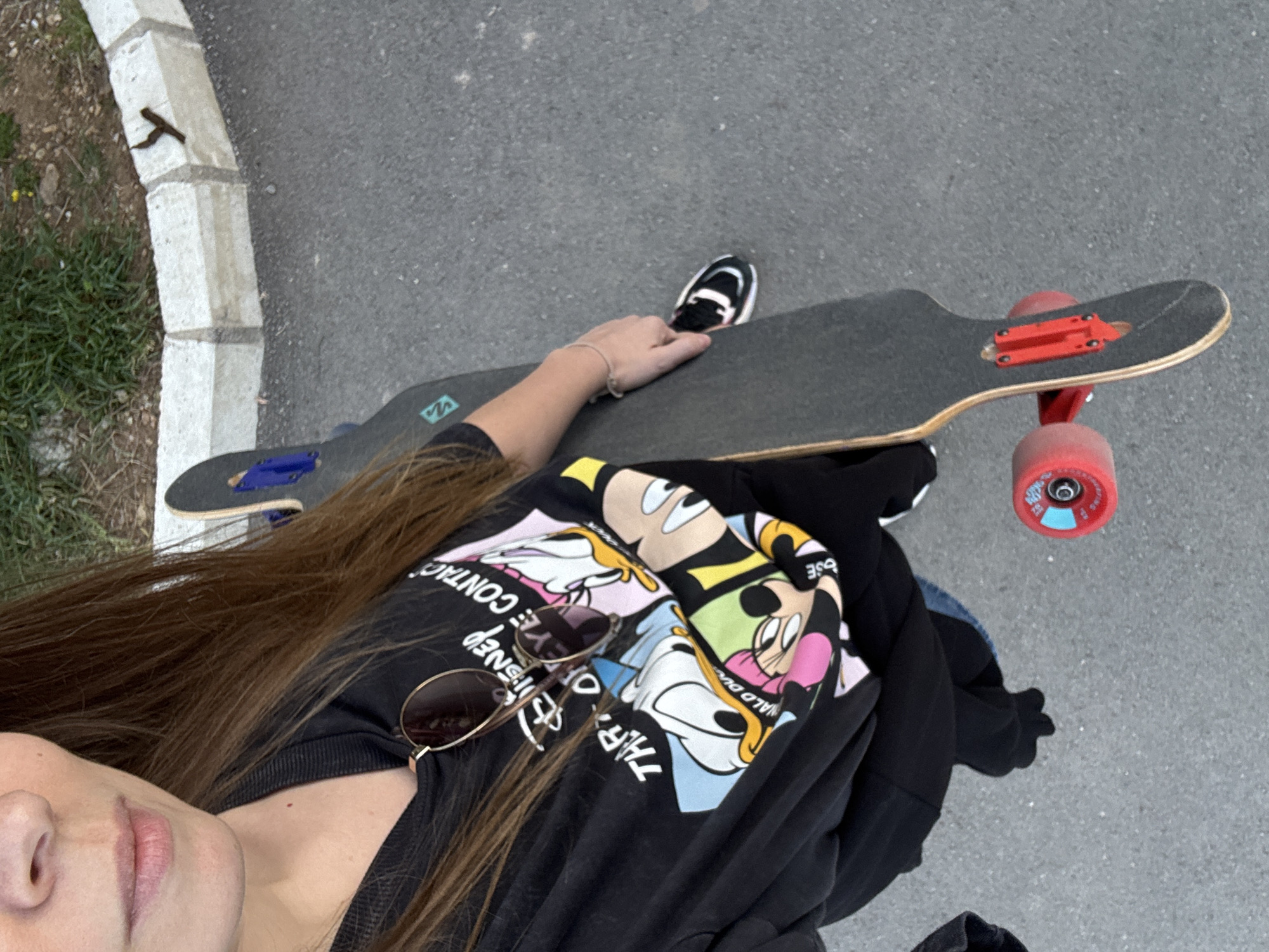 The image presents a first-person perspective of a person with long brown hair and a partial view of their face (chin and mouth), positioned alongside a longboard skateboard on an asphalt surface. The setting is outdoors in Budva, Montenegro, next to a concrete curb with some grass and dirt visible beyond it.
The individual is wearing a black t-shirt adorned with colorful cartoon panels featuring classic Disney characters such as Mickey Mouse, Minnie Mouse, Donald Duck, and Goofy. A pair of sunglasses with thin metal frames rests on the collar of their shirt. Their right hand, wearing a simple bracelet, is placed on the black grip tape of the longboard.
The longboard features red trucks and bright red wheels, with one wheel partially showing the text "ROCKERS 72MM 82A". A light blue logo, possibly "VZ", is visible on the underside of the board near the front trucks. The lighting indicates it is daytime, likely sunny.
Fragments of text are visible on the t-shirt, appearing inverted and partially obscured: "DISNEY", "EYE CONTA", "THR", and "ALD DU".