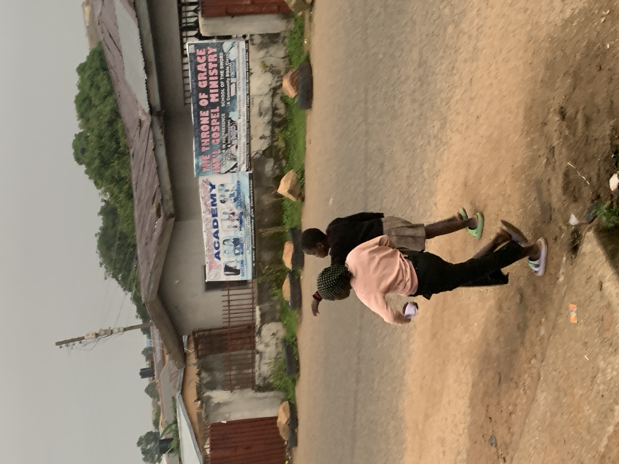 The image depicts two young children walking on a dirt road in Jos, Nigeria. The child in the foreground, on the left, is wearing a light pink long-sleeved shirt, black pants, and white and gray sandals. This child is holding a white object in their right hand. The child behind them, on the right, is wearing a dark long-sleeved shirt and light-colored shorts, with green sandals. This child is pointing towards the left.
In the background, there is a building with a corrugated metal roof. A banner is prominently displayed on the building, advertising "THE THRONE OF GRACE INT'L GOSPEL MINISTRY" and "PRECIOUS ACADEMY". The banner also lists services such as "DAYCARE", "NURSERY", "PRIMARY", "SECONDARY", "AN HOUR IN HIS PRESENCE (Prayer & Intercession)", and "SCHOOL OF THE SWORD (A Community Bible Class)". The general environment appears to be a somewhat unkempt urban or semi-urban area, with electrical poles and wires visible. The road surface is a mix of dirt and some scattered debris. The lighting suggests an overcast or cloudy day.