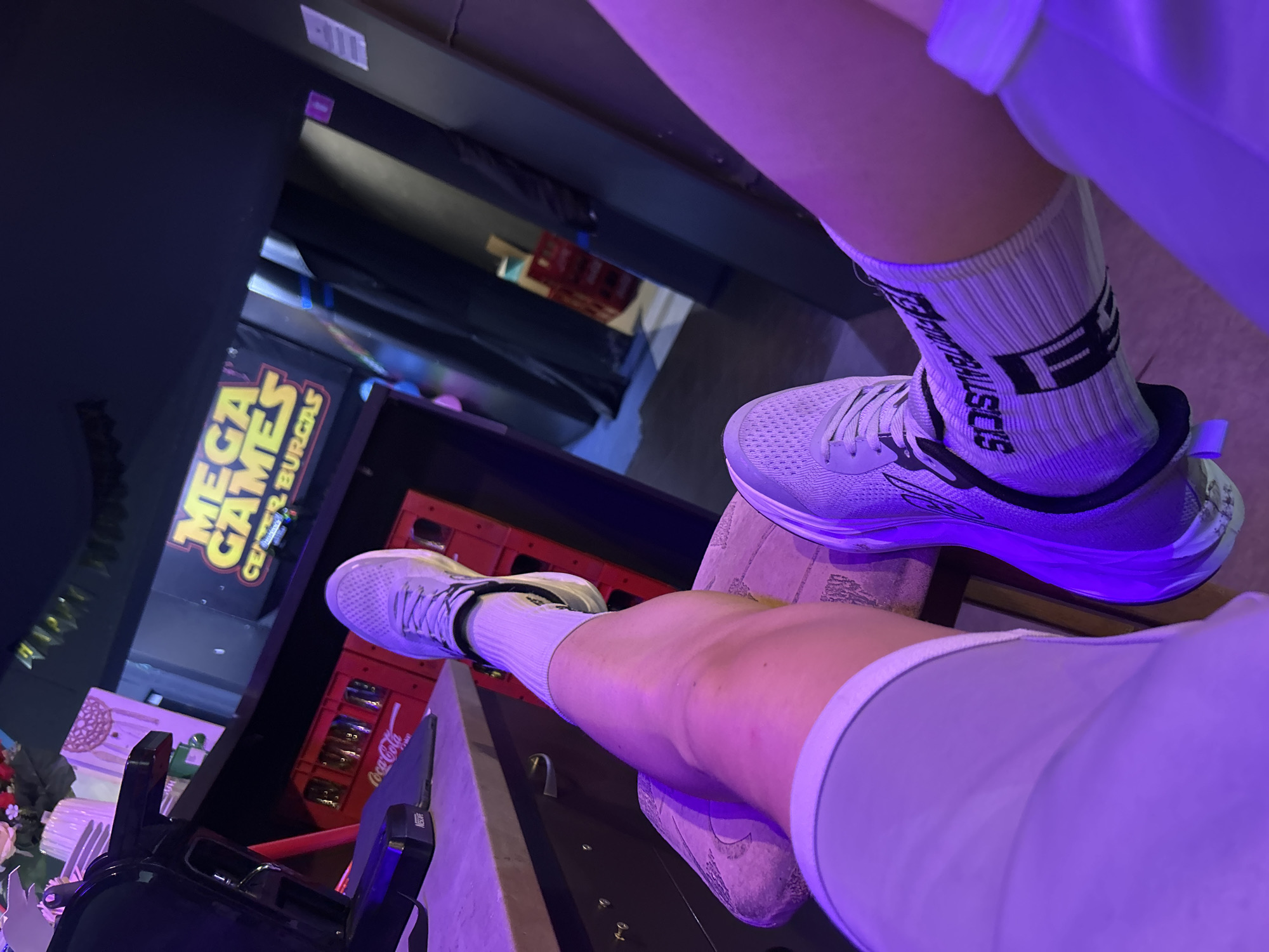 The image captures a casual indoor setting, likely an entertainment venue or restaurant, characterized by dim, purplish ambient lighting. The perspective is a low-angle shot looking upwards, focusing on two pairs of legs belonging to two individuals, possibly resting or lounging.
Both individuals are wearing white athletic shoes and white, ribbed athletic socks. One sock, visible on the right leg, displays stylized dark lettering that appears to read "SISTIMUSCLES" and "FE". The legs are propped up; one is positioned to the left and lower, while the other is elevated on the right side of the frame. Both appear to be wearing light-colored shorts or athletic wear.
In the background, a large sign with prominent yellow lettering reads "MEGA GAMES" above "CENTER BURG...", suggesting an arcade or gaming center, possibly offering food. This sign provides a strong location cue, indicating Burgas, Bulgaria, as the setting. Below the sign, several red crates are stacked, one of which clearly displays "Coca-Cola". To the far right, a small black appliance with the word "Nescafe" is visible. Other background elements are less distinct due to the low light, but include dark furniture or counters and what might be artificial flowers on a table.
The atmosphere is relaxed, with the individuals' legs propped up, suggesting they are taking a break or enjoying their time in the establishment. The artificial purple lighting indicates an evening or nighttime setting.