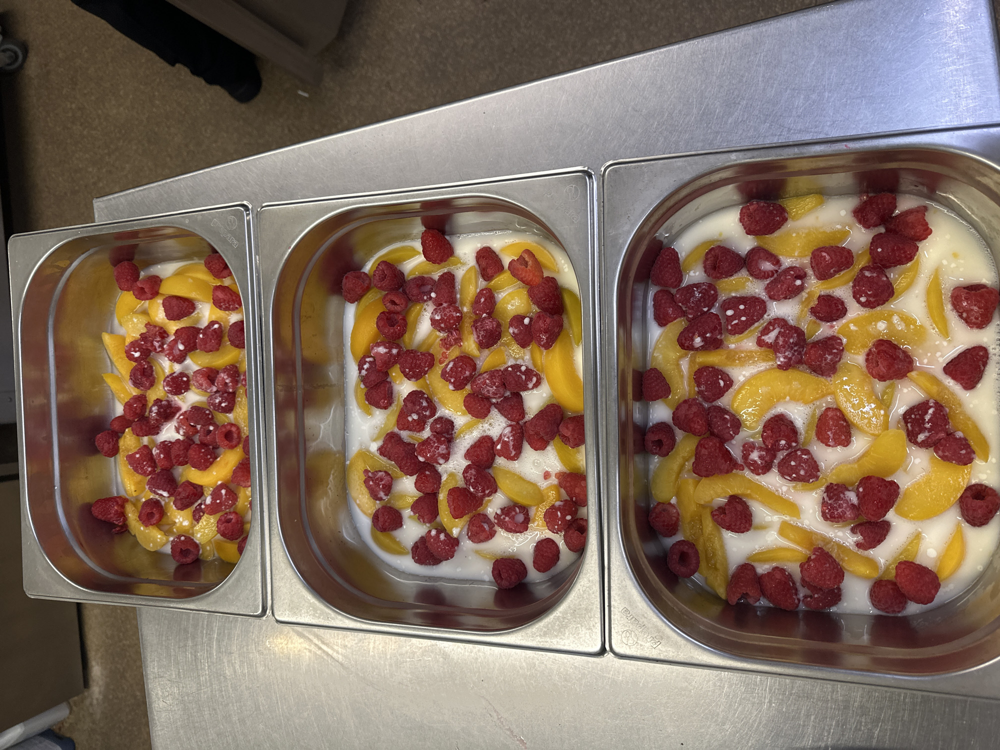 The image shows three stainless steel food pans filled with sliced peaches and raspberries in a creamy white liquid, likely a dessert or compote. The pans are arranged side-by-side on a stainless steel surface, suggesting a commercial kitchen or food preparation area. The lighting is bright and overhead, indicating an indoor setting. The food appears fresh, with the raspberries exhibiting their characteristic texture and bright red color, and the peaches are a vibrant yellow-orange. The creamy liquid has small white flecks, possibly indicating dairy or a thickening agent. The overall scene is one of food preparation, with the focus on the appetizing ingredients. There is no visible text in the image.
