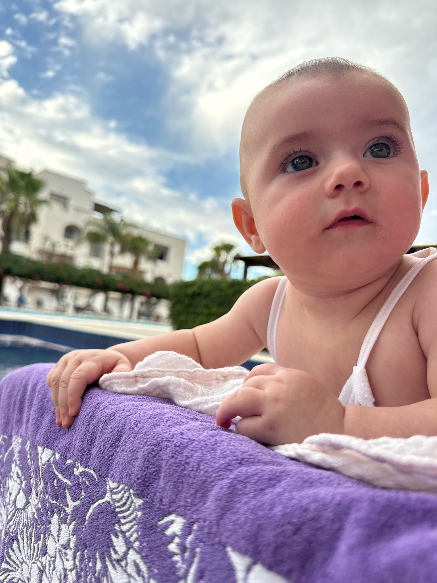 A close-up, low-angle shot shows a baby resting on a purple towel with a white towel draped over it. The baby is wearing a white garment with thin straps. In the background, out of focus, is a swimming pool, a building with arched windows, palm trees, and a cloudy blue sky. The scene appears to be outdoors, likely at a resort or hotel in Isola delle Femmine, Italy, given the architecture and vegetation.