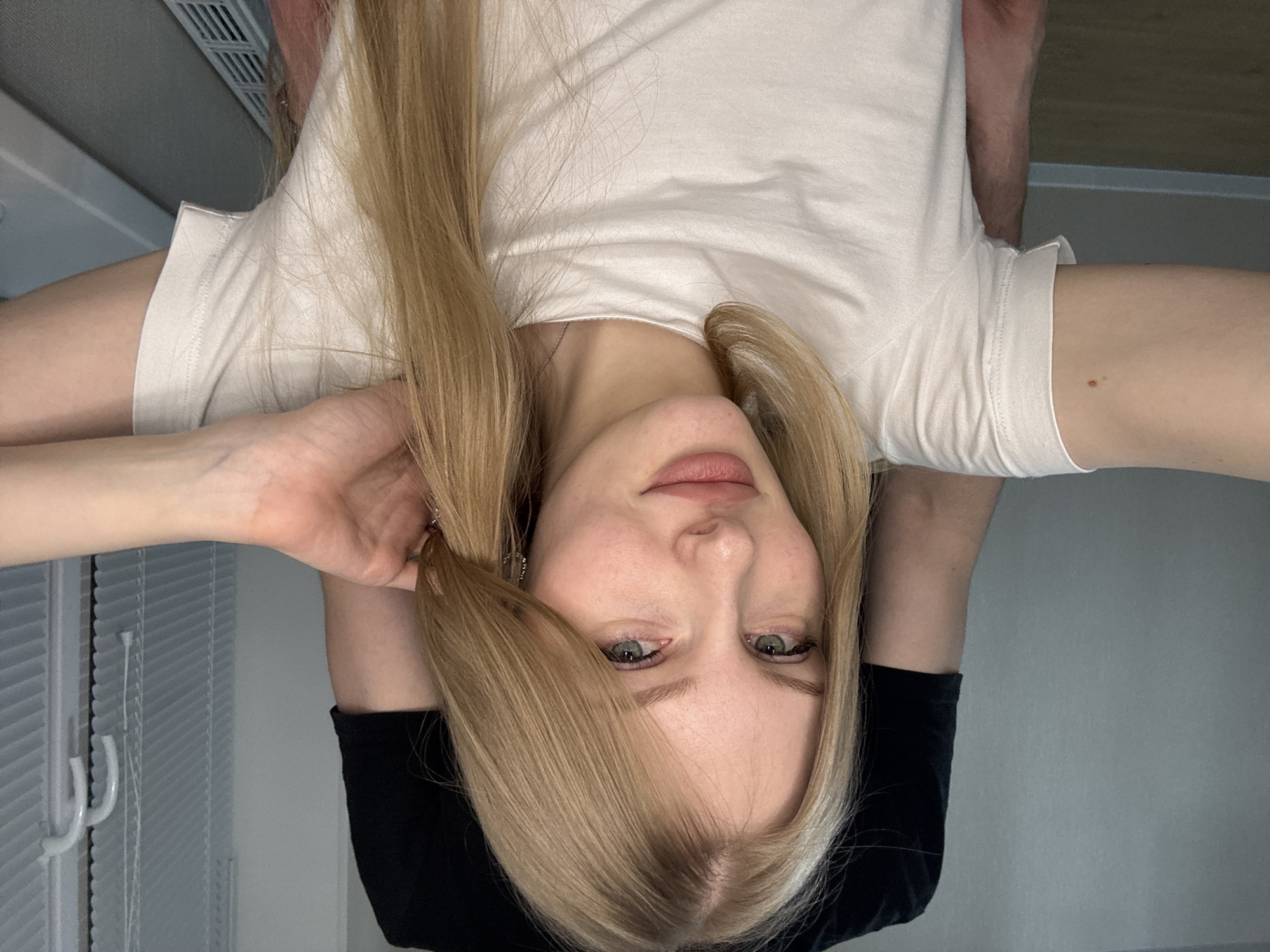 The image features a young woman with long, straight blonde hair, presented in an inverted orientation, suggesting she is lying on her back or posing upside down. Her head is centered, and she looks directly at the viewer with light-colored eyes and a neutral, slightly contemplative expression. Her lips are softly parted.
She is dressed in a plain white short-sleeved t-shirt. Her right hand (from the viewer's perspective, her left hand) is raised and gently touching her blonde hair near her ear. Her left arm (viewer's right) extends outwards, displaying fair skin with a small, dark mole on the upper part. A black fabric or garment is visible underneath her head and extending down her left arm, possibly part of her clothing or a surface she is resting on.
The background on the left side of the image reveals white or light-colored horizontal blinds, indicating an indoor setting. On the right, the background is a smooth, plain, light grey wall. The scene is well-lit, characteristic of daytime or bright interior lighting. The photograph, taken in Perm, Russia, captures a moment of calm. There is no visible text.