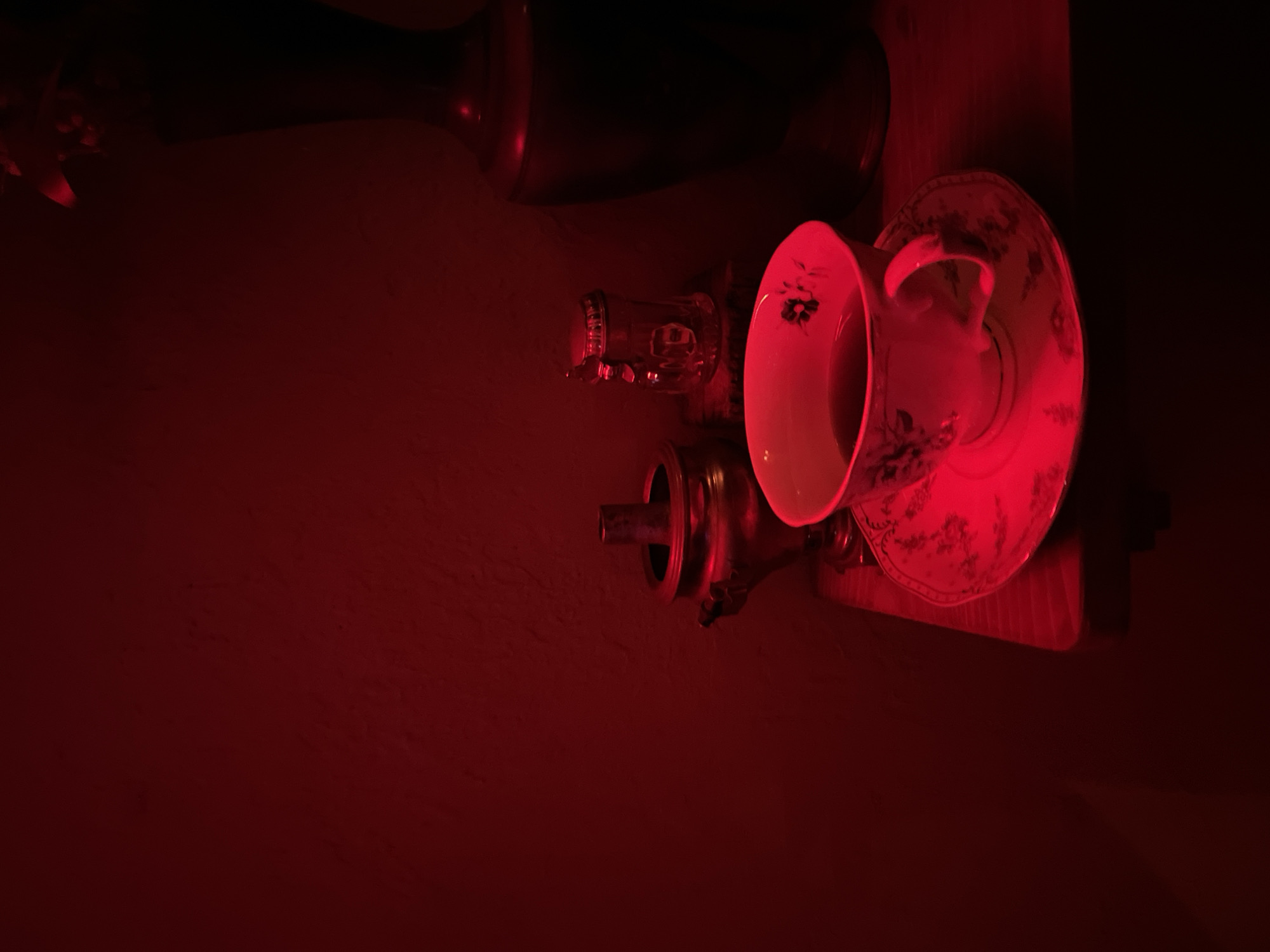 The image, bathed in a red light, presents a still life scene. A patterned teacup sits on a matching saucer, next to a glass. Below them is an ornate metal teapot or beverage dispenser. Above these items is a vase. The objects are arranged on a wooden shelf. It is unknown whether this scene is set in Tbilisi, Georgia.