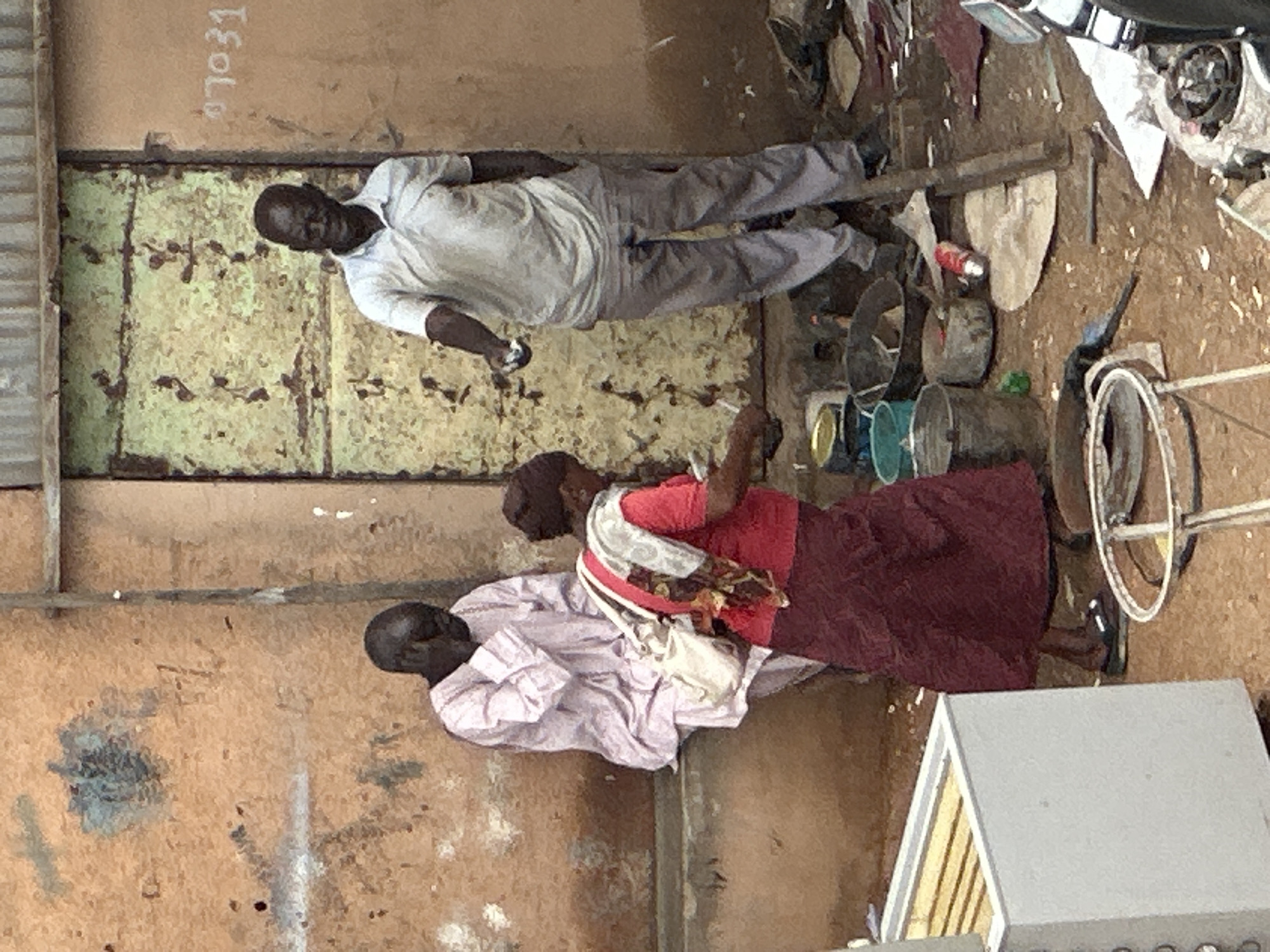 In Jos, Nigeria, an outdoor scene depicts three individuals near a dilapidated building entrance. On the left, a person in a light purple traditional garment sits on a concrete ledge, with their head bowed and hand covering their face. In the center, a person wearing a red shirt and a dark maroon skirt is partially visible, facing away from the camera and appearing to interact with something on the ground. To the right, a man in a light-colored polo shirt and grey trousers stands, facing the camera and extending his right hand, holding a small silver object.
The environment is cluttered with various objects, including buckets, pots, debris, and a white rectangular structure that resembles a refrigerator or display case on the far left. A worn, green metal door is visible behind the standing man. Graffiti or markings are present on the building's exterior walls, including the number "07031" in white. The ground is a dirt and gravel surface. The overall impression is of an informal marketplace or an area with limited resources.