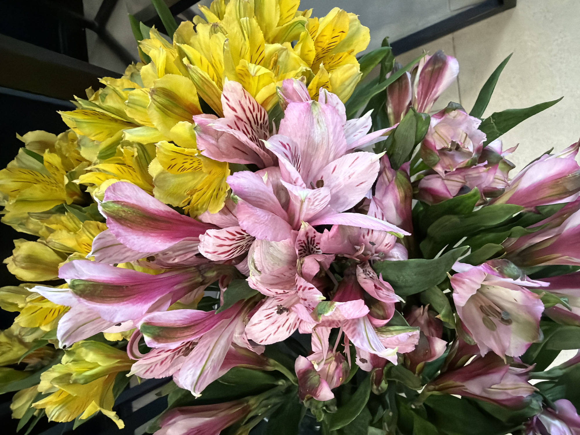 A close-up, overhead shot shows a vibrant bouquet of pink and yellow alstroemeria flowers. The flowers are arranged loosely, with some of the pink blooms in the foreground showing delicate stripes and a hint of darker veins. The yellow flowers, in the background, are fully open and bright. Green leaves are interspersed throughout the bouquet. The background is dark and out of focus, with hints of a dark metal structure on the right. The scene appears to be indoors, possibly in a flower shop or home, with a light-colored floor visible at the bottom right corner.