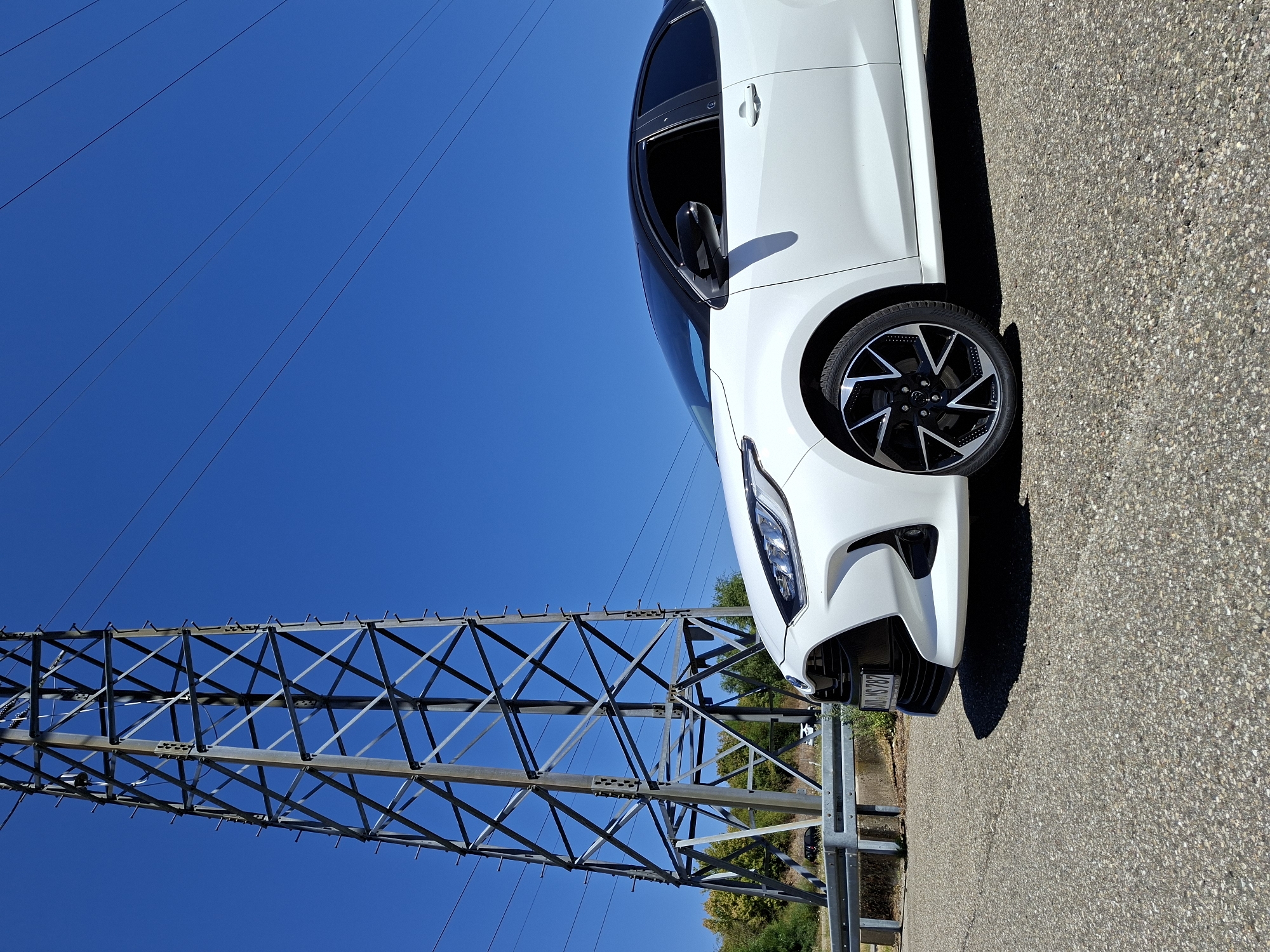 The image shows a white Toyota Yaris with a black roof parked on an asphalt road under a clear blue sky. To the left of the car is a large, grey metal electricity pylon. In the background, there are trees and a roadside barrier. The license plate of the car reads "BAD MS 787." This license plate format is consistent with registration plates issued in the district of Rastatt (associated with Baden-Baden, Germany), indicating the car is likely located in or around Baden-Baden. There are no people visible in the image.