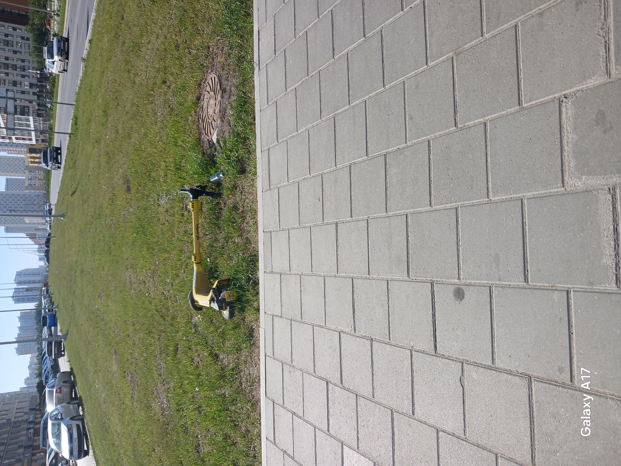 A ground-level, outdoor photograph depicts a sidewalk constructed of gray paving stones on the right, bordered by a grassy area on the left. In the background, cars are parked alongside a road, and taller buildings are visible in the distance, which indicates a city setting.
Specific objects in the scene include a manhole cover and a yellow electric scooter parked in the grass. The scooter has visible license plates.
The metadata indicates the image was taken using a Galaxy A17 mobile device. Given the prompt, the location is specified to be in the city of Yekaterinburg, Russia.
