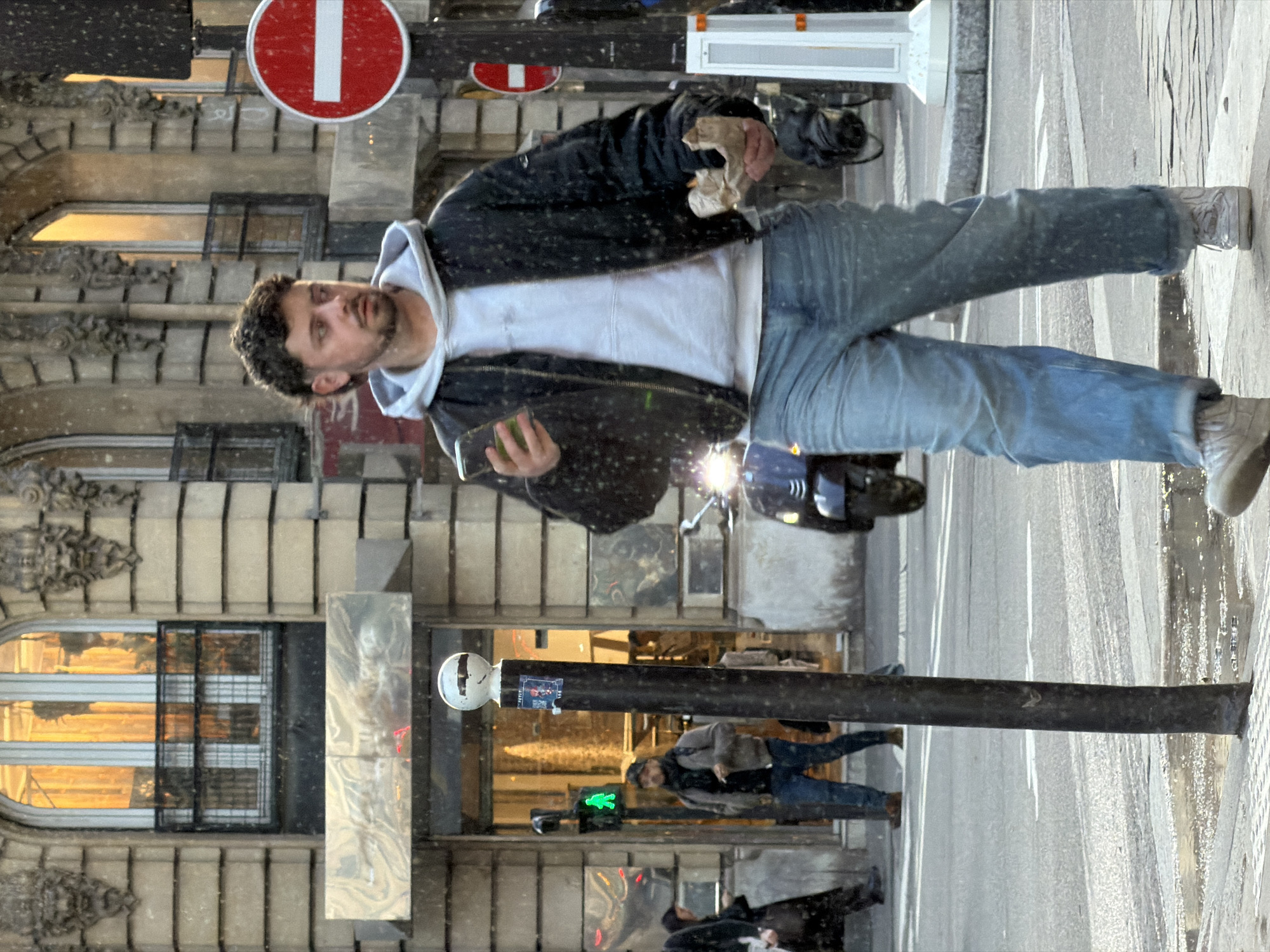The image presents an urban street scene in Paris, France, under conditions of light precipitation, likely snow or sleet, indicated by white specks in the air and on surfaces.
In the foreground, a male, approximately 25-35 years old, with dark curly hair and a short beard, walks on the sidewalk. He wears a light grey hooded sweatshirt under a black bomber jacket, light blue jeans, and light-colored sneakers. His clothing is visibly speckled with white precipitation. He looks towards his right, slightly away from the camera. He holds a light-colored smartphone in his left hand and a crumpled brown paper bag in his right hand.
To the left of the main subject, a partially obscured individual in dark attire walks away. Further back, another male figure in a dark jacket and jeans walks past a building entrance, positioned near a green pedestrian signal displaying a walking figure. A dark-colored scooter or moped with an illuminated headlight is visible in the middle ground, to the right of a black street pole topped with a white sphere.
The background features a multi-story building with a light-colored, ornate stone facade, consistent with Haussmannian architecture. It displays large arched windows with white frames, small black metal balconies, and decorative carvings above the windows. The ground floor incorporates modern, reflective silver panels above storefront entries.
Street infrastructure includes a prominent black pole with a white spherical top in the immediate foreground, and a white pole to the right displaying a red circular "No Entry" (sens interdit) traffic sign. The pavement and street are wet, with puddles present on the sidewalk and at the road's edge. White pedestrian crossing lines are visible on the asphalt street surface.