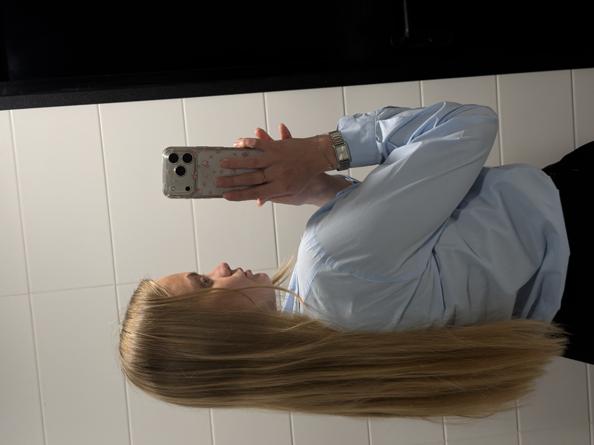 A young woman with long blonde hair is holding a cell phone. She is wearing a light blue button-down shirt and a watch on her left wrist. The background appears to be a bathroom setting, with white tiled walls. There is no information to suggest the image was taken in Izhevsk, Russia.