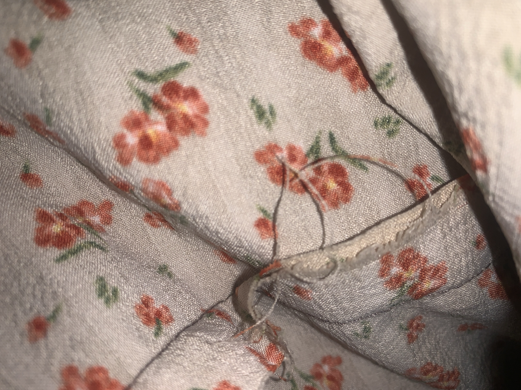 The image shows a close-up view of a light-colored fabric with a floral print of small red flowers and green leaves. The fabric appears to be wrinkled and folded, with a seam and some loose threads visible in the center of the image. The lighting is soft and casts shadows, highlighting the texture of the fabric. There are no people or any specific identifiable landmarks visible that would allow for the determination of a location in Jauro Yinu, Nigeria. The image is a still photograph of textile material.