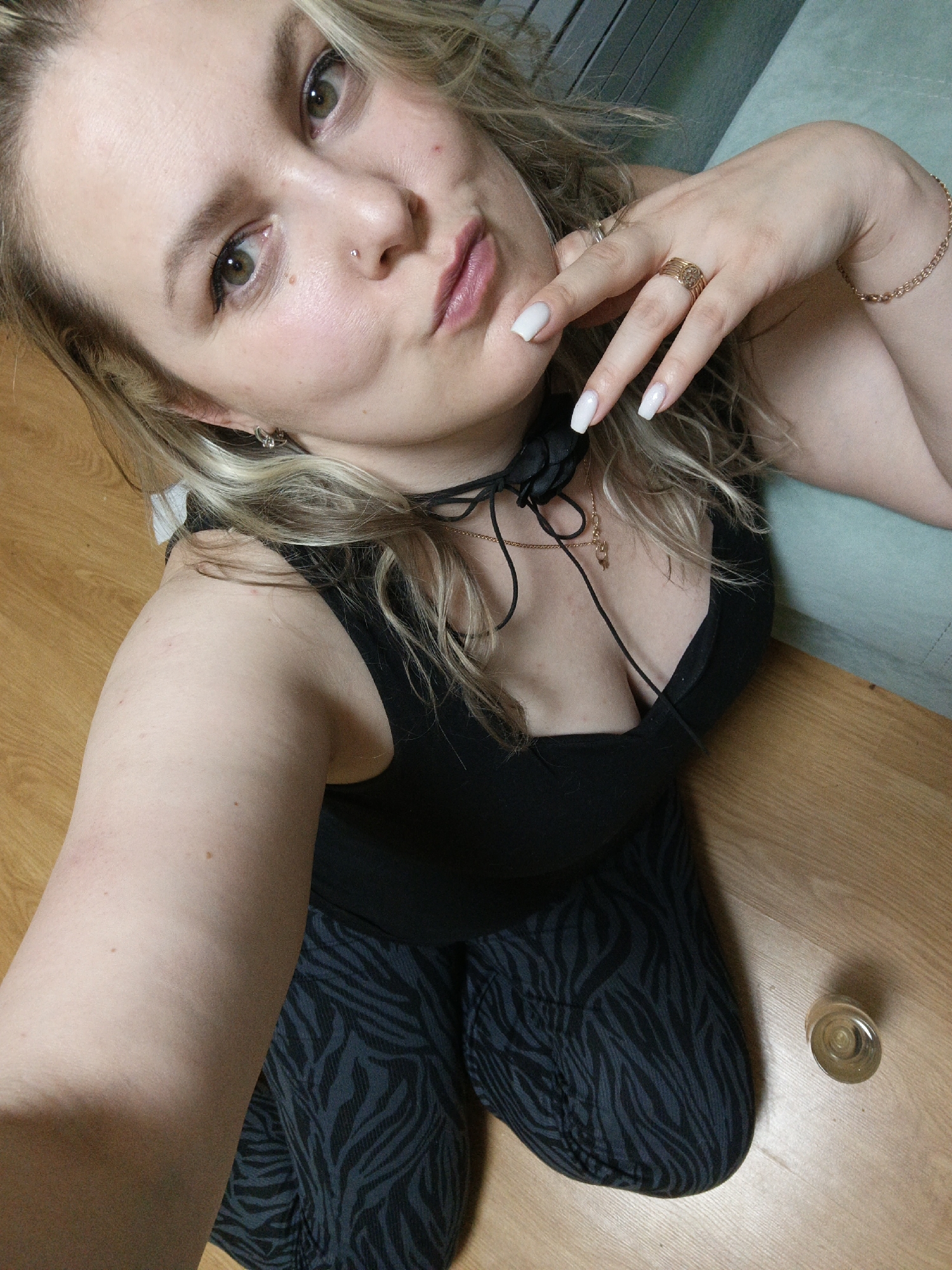 A woman is posing for a selfie. She has blonde hair and is wearing a black tank top and patterned leggings. She is kneeling on a wooden floor. Her hand is raised to her face, showcasing a ring and bracelet. A small glass object is visible on the floor near her legs. A portion of a sofa is visible in the background. There is no discernible evidence to indicate that this photo was taken in the city of Chaikovsky, Russia.