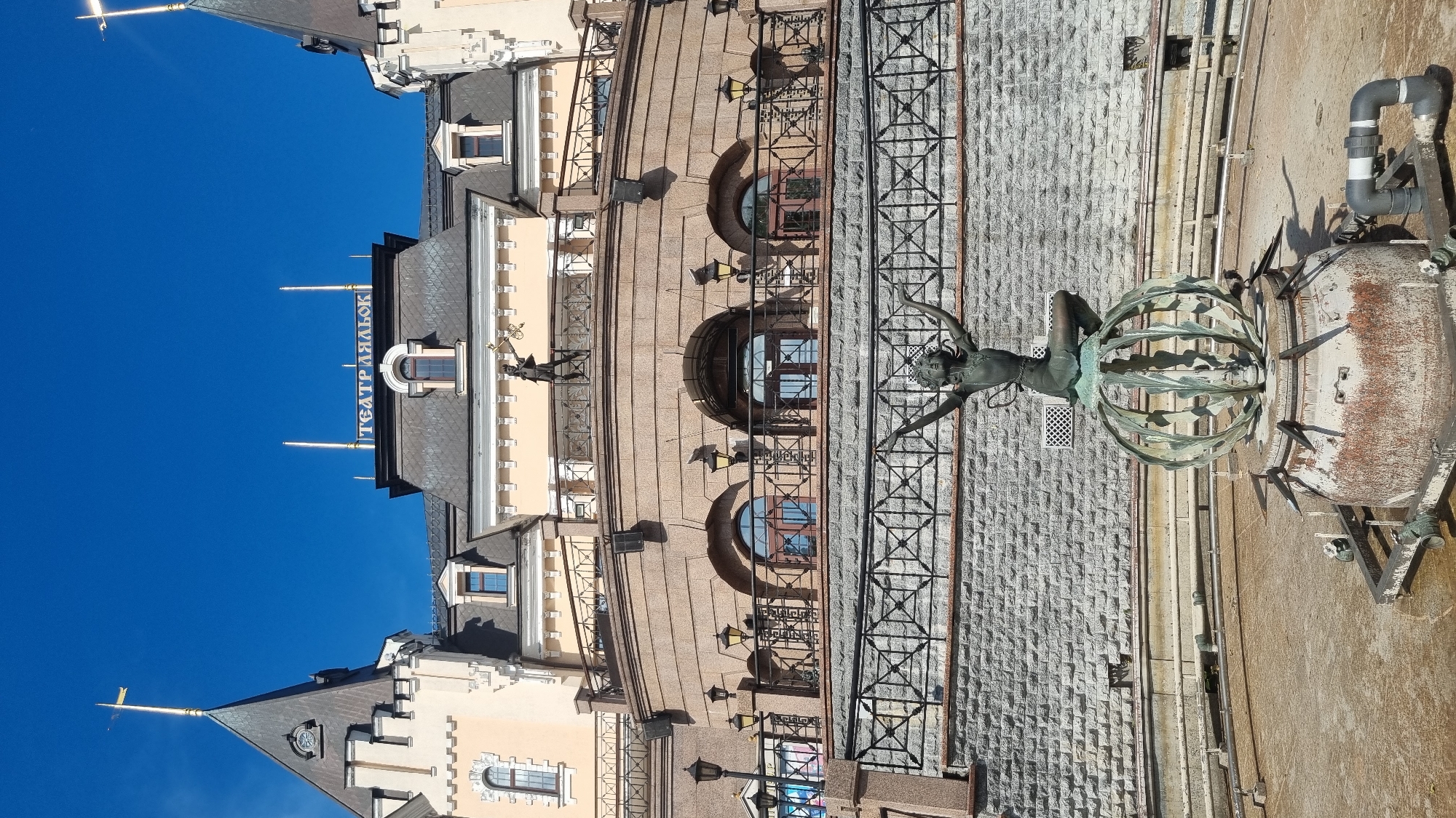 This image captures a close-up view of the façade of a building, likely a theater, with a clear blue sky in the background. The building's architecture is ornate, featuring multiple levels, arched windows, decorative railings, and a prominent sign with Cyrillic text. A bronze statue of a woman perched on a spherical, cage-like structure is a central element, positioned in front of the building's stone-clad lower section. The statue appears weathered, suggesting it is an older piece. The lighting indicates it is daytime, likely morning or afternoon, with distinct shadows cast by the building and the statue. The overall scene is static, focusing on the architectural details and the sculptural element, possibly a fountain or part of a historical monument. The visible text reads "ТЕАТР ЯЛЬОК," which translates to "Puppet Theatre" in Ukrainian, confirming the building's purpose. The scene is set outdoors, with the bright sky and clear visibility suggesting fair weather.