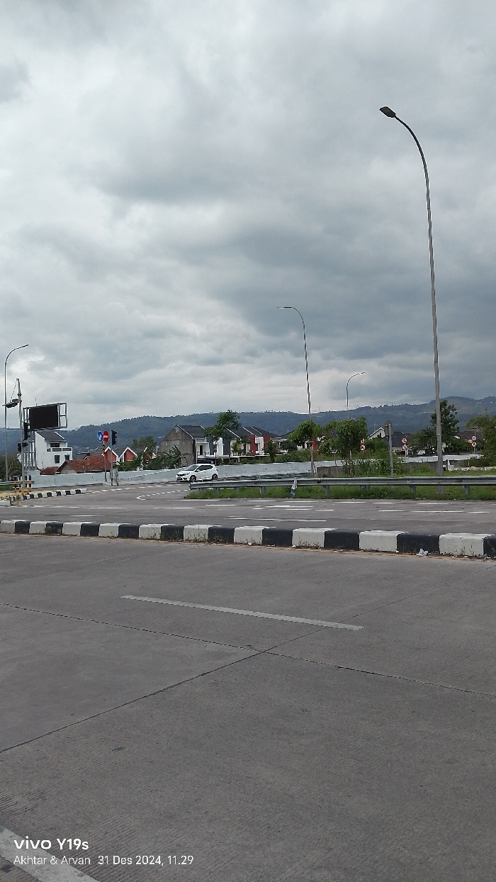 An outdoor, eye-level shot captures a road intersection or merge point in Sumedang, Indonesia, under an overcast sky. In the foreground, a wide asphalt road stretches across the bottom of the frame, featuring white road markings and a concrete median barrier with alternating black and white blocks.
In the midground, a white compact car is visible on a road that appears to curve to the left, possibly at a junction. To the left, a traffic light pole with a black rectangular structure, possibly a billboard or digital sign, stands next to a white building. Several streetlights with tall, curved poles are distributed throughout the scene, reaching towards the sky. A metal guardrail runs along the right side of the road, separating it from an elevated area.
The background reveals a residential area with multiple houses, some featuring red roofs and others predominantly white, built on gentle slopes. Behind the houses, green, undulating hills extend into the distance, partially obscured by the heavy cloud cover. The sky is uniformly grey and cloudy, indicating an overcast day.
A watermark in the bottom left corner displays "vivo Y19s", "Akhtar & Arvan", and the date and time "31 Des 2024, 11.29". The scene appears quiet with minimal traffic.