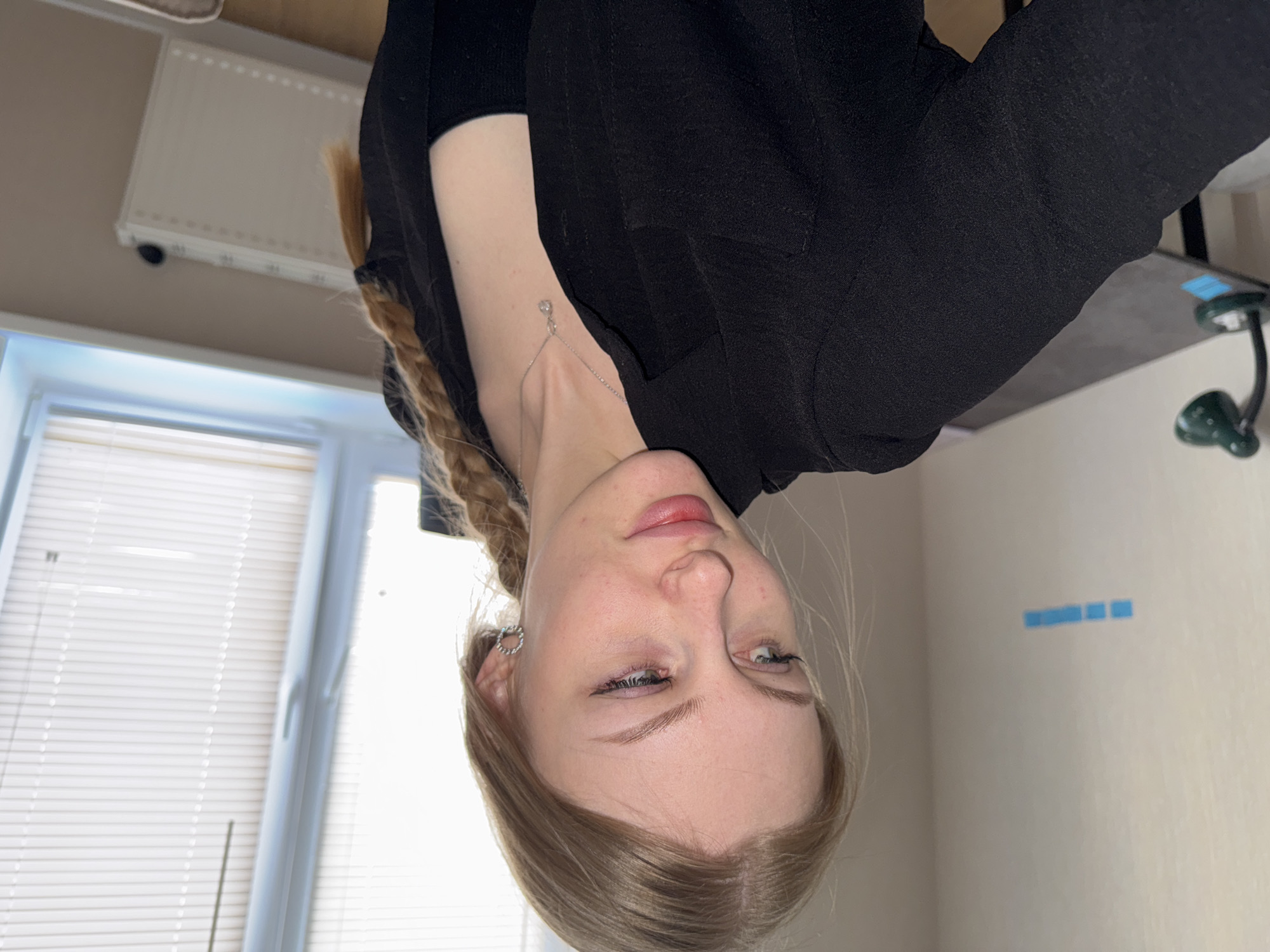 A fair-skinned woman with blonde hair is oriented upside down in the frame. Her hair is braided and styled to the side. She wears a black garment that appears to be a blazer. A delicate silver necklace is visible on her neck. In the background, there's a window with horizontal blinds, a white radiator, a desk with a green lamp, and a plain wall. The blue squares on the wall are not identified. There is no explicit information in the image that confirms the location is Perm, Russia.