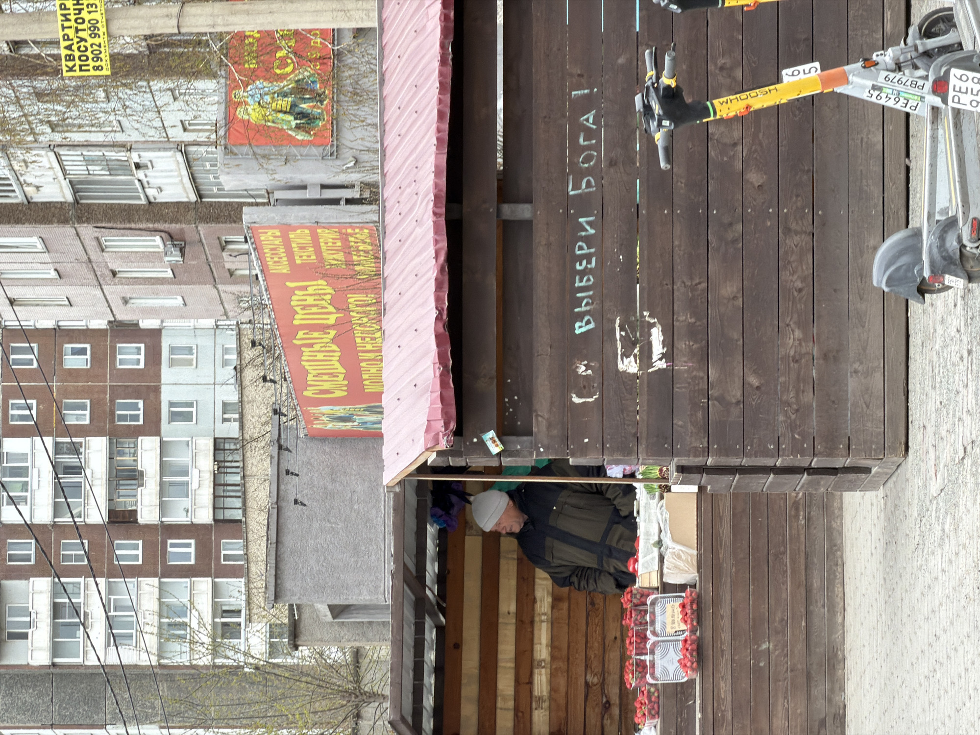 The image shows a street scene in Krasnoyarsk, Russia. A man in a grey hat and dark jacket is standing inside a small wooden kiosk, possibly a fruit or vegetable stand, as trays of red berries are visible on the counter. The kiosk has a red roof and a sign with the words "ВЫБЕРИ БОГА!" (Choose God!) written on the exterior wall.
Two parked electric scooters with yellow accents and "WHOOSH" branding are positioned near the kiosk. One scooter's license plate number is "PE6495" and the other is "PB7979."
Across the street, an apartment building stands tall, its windows showing signs of age and wear. On the side of the building, a large advertisement is visible. A sign reading "СМЕШНЫЕ ЦЕНЫ" (Funny Prices) is also seen. Overhead power lines run in front of the building.