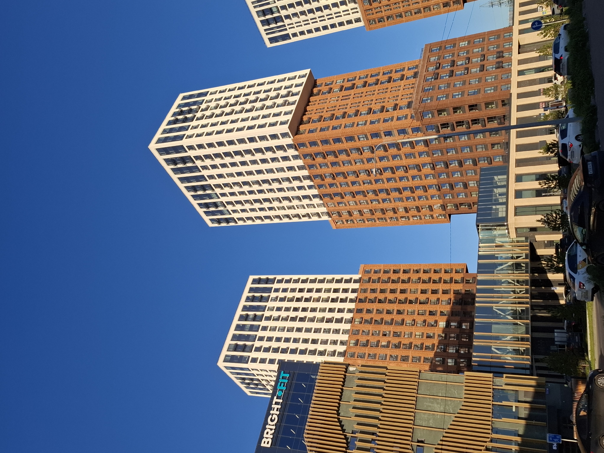 The image shows a modern architectural complex with multiple buildings against a clear blue sky. The buildings are tall and feature a mix of materials, including brick and white facades with numerous windows. One building has the text "BRIGHT FIT" on its dark blue facade. The buildings are arranged in a staggered manner, with some appearing to be perched on top of others. The lower sections of some buildings have decorative wooden elements and glass. In the foreground, there is a street with several cars parked and driving. The environment appears to be an urban setting, likely a city, bathed in sunlight. There are no people clearly visible in the image.