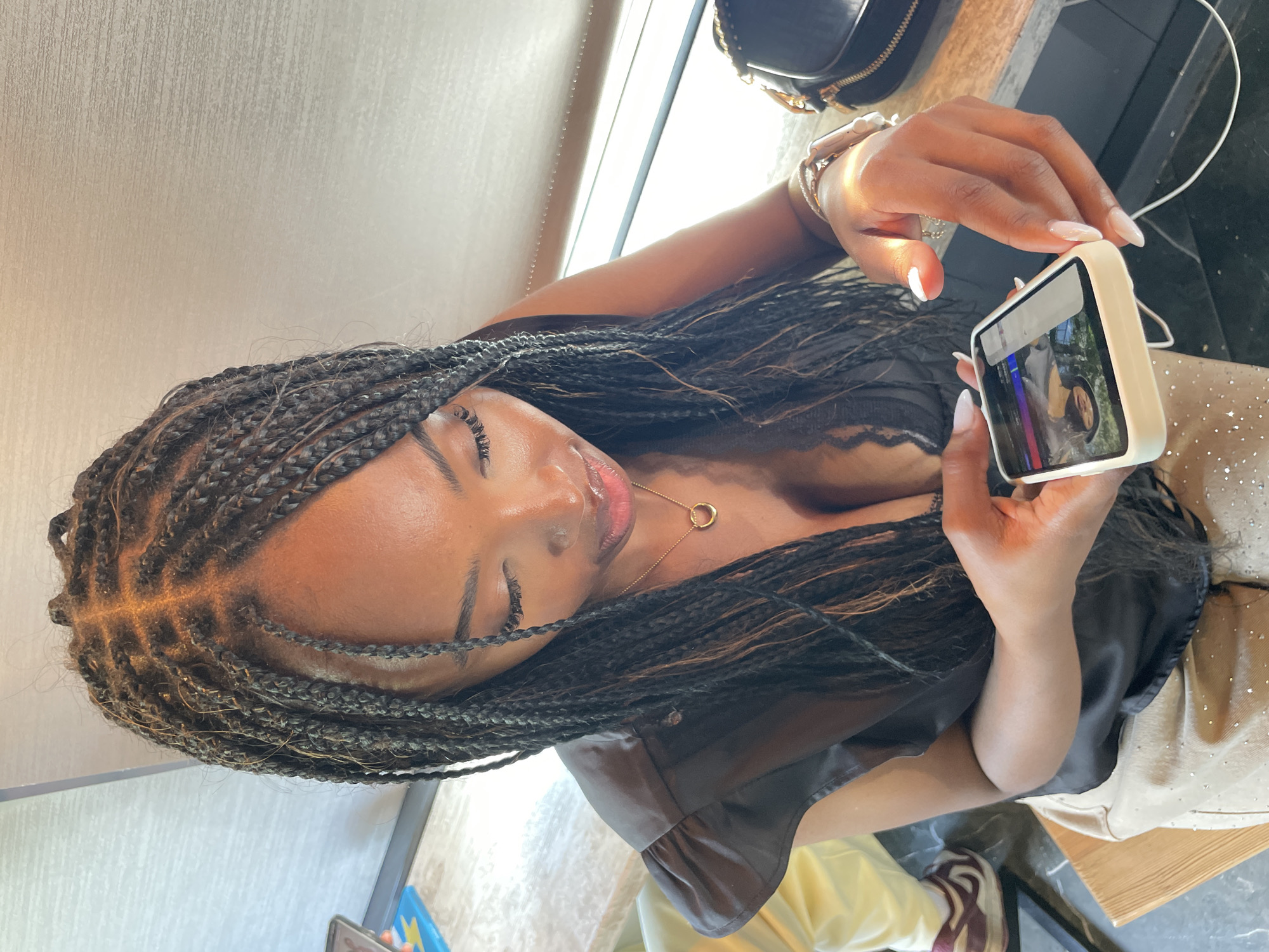 A dark-skinned woman with long, braided hair is sitting at a table, looking down at her phone. She is wearing a black top and light-colored pants with a sparkly embellishment. The phone is white with a light-colored case. The background shows a window with blinds and a black purse on the table. The scene appears to be indoors.