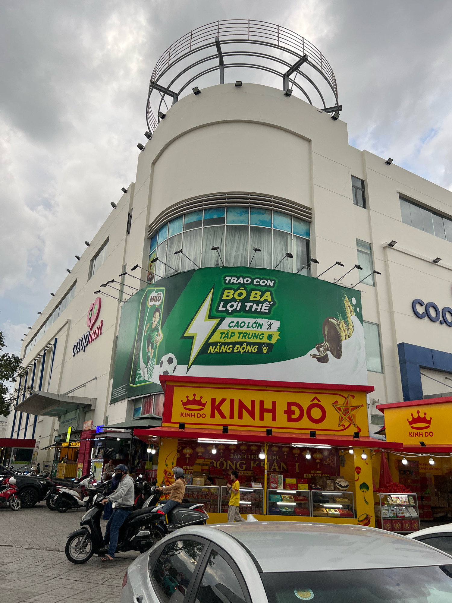 The image depicts a street scene in front of a Co.opmart supermarket in Nha Trang, Vietnam. A large, modern building with a distinctive curved top and a circular balcony railing dominates the upper portion of the frame. Several billboards are mounted on the building's facade. One prominent green billboard features an advertisement for Milo, showing a young boy playing soccer and text in Vietnamese. Below this, a bright yellow stall with red trim displays the "Kinh Do" brand, suggesting a bakery or food retailer. Several people are visible, some on motorcycles in the foreground and others walking or working at the yellow stall. A silver car is partially visible in the lower foreground. The sky is overcast, indicating a cloudy day. The overall scene is busy and typical of a commercial area in Vietnam.
