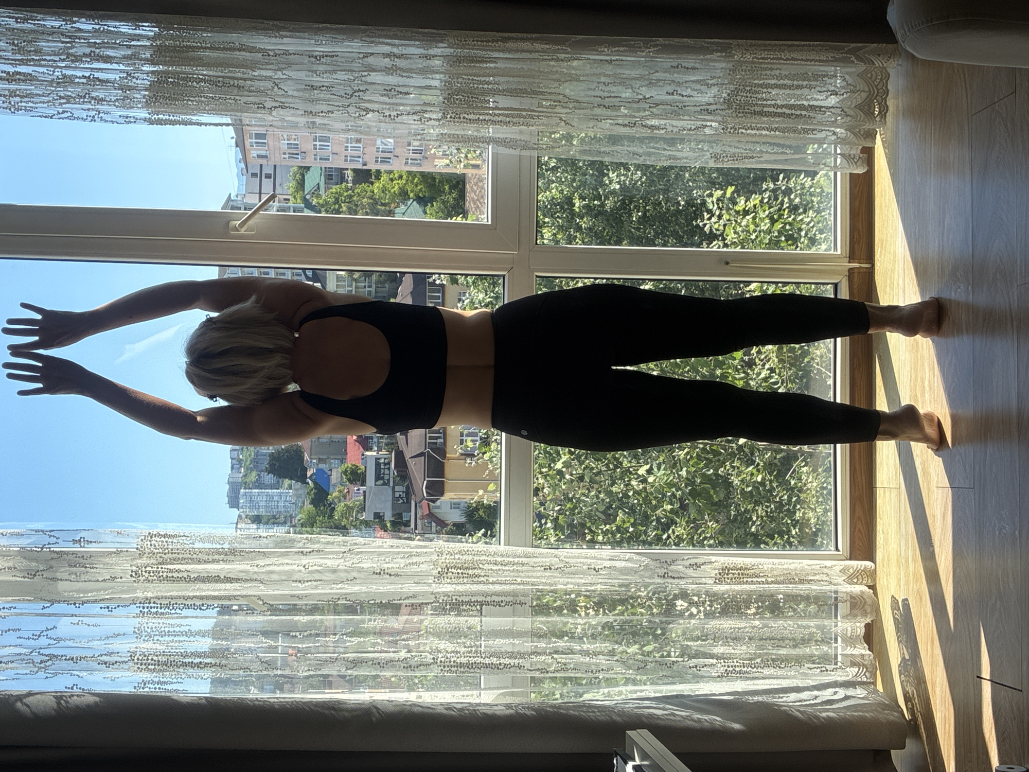 The image depicts a woman with blonde hair, viewed from behind, performing a yoga pose with her arms raised and hands touching the top of a large window frame. She is wearing a black sports bra and black leggings, standing on a wooden floor. Sunlight streams through the window, casting shadows on the floor. The window overlooks a cityscape with buildings and lush green trees, suggesting an urban environment in Sochi, Russia. The window is adorned with sheer white curtains with a decorative pattern. The scene appears to be an indoor space, likely a room with a view.