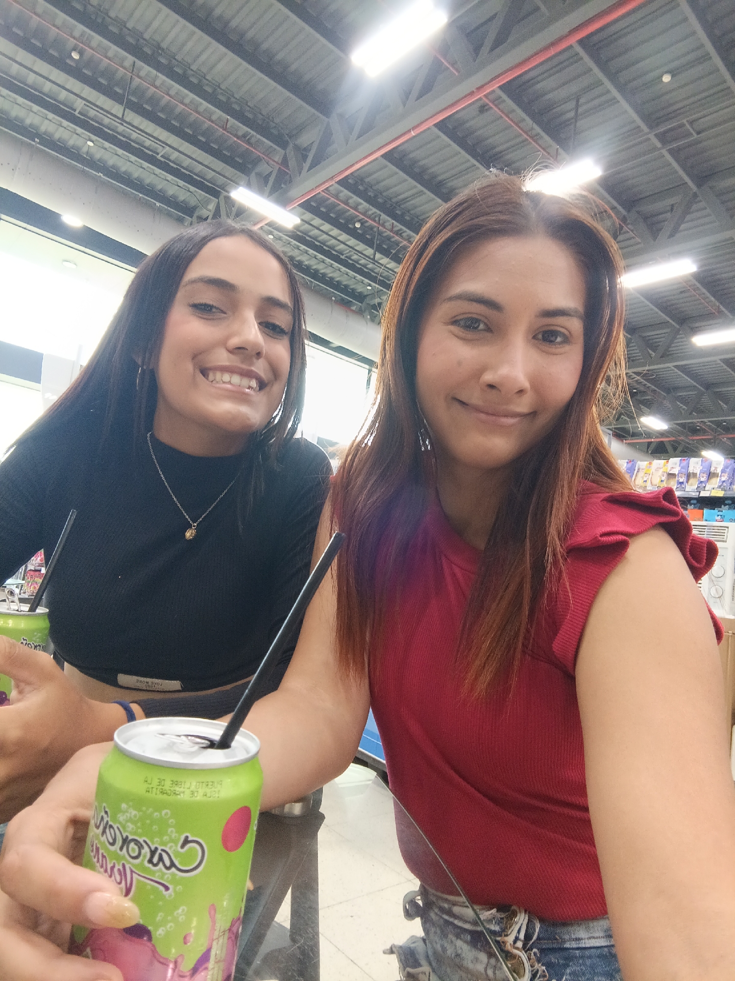 Two women are seated at a table indoors. The woman on the left has long, dark hair, wears a black top and a necklace. She is smiling. The woman on the right has shoulder-length brown hair and wears a red top. She is also smiling. Both have a beverage in a green can with a black straw. The background includes an industrial-style ceiling, possible shelves, and merchandise. The location appears to be a market or store, but not specifically identifiable as Juan Griego, Venezuela without further details.