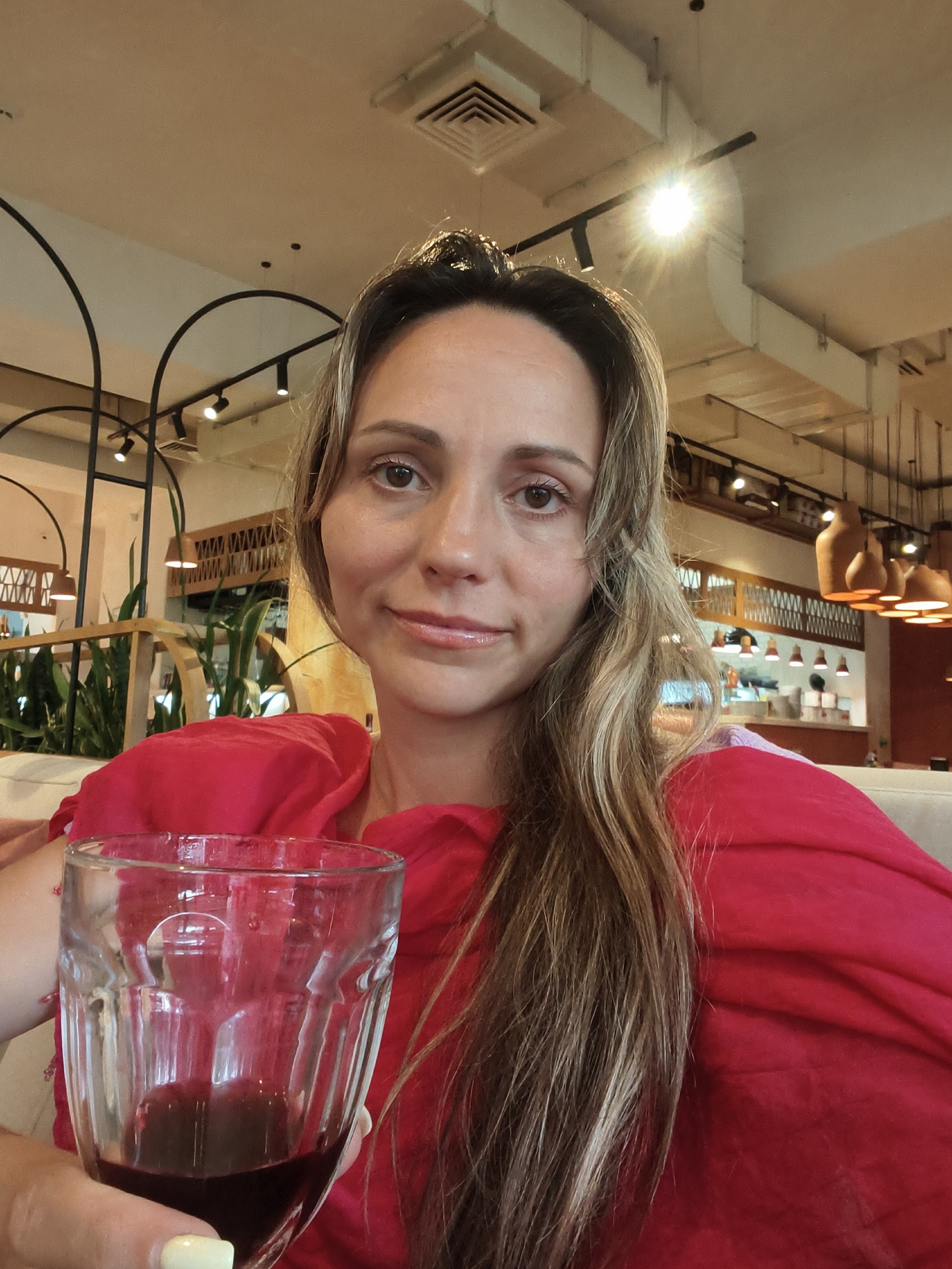 The image shows a selfie of a woman holding a glass of red liquid, presumably wine. She is wearing a vibrant red top and has long, blonde hair. She is sitting indoors, possibly in a restaurant or cafe, as indicated by the background which shows interior lighting, architectural details, and plants. The scene is well-lit, with a bright light source visible in the upper right quadrant of the image. The woman is looking directly at the camera with a slight smile. The overall impression is of a relaxed and informal moment. The location is identified as Sochi, Russia, suggesting the woman is at a venue within that city.