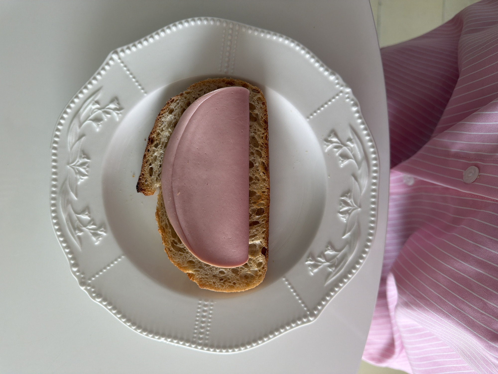 A top-down shot shows a white decorative plate on a white table. A piece of bread sits on the plate, topped with a slice of processed meat. To the right of the plate, a pink and white striped shirt is partially visible. No specific location in Khimki, Russia, can be inferred from the visual information provided.