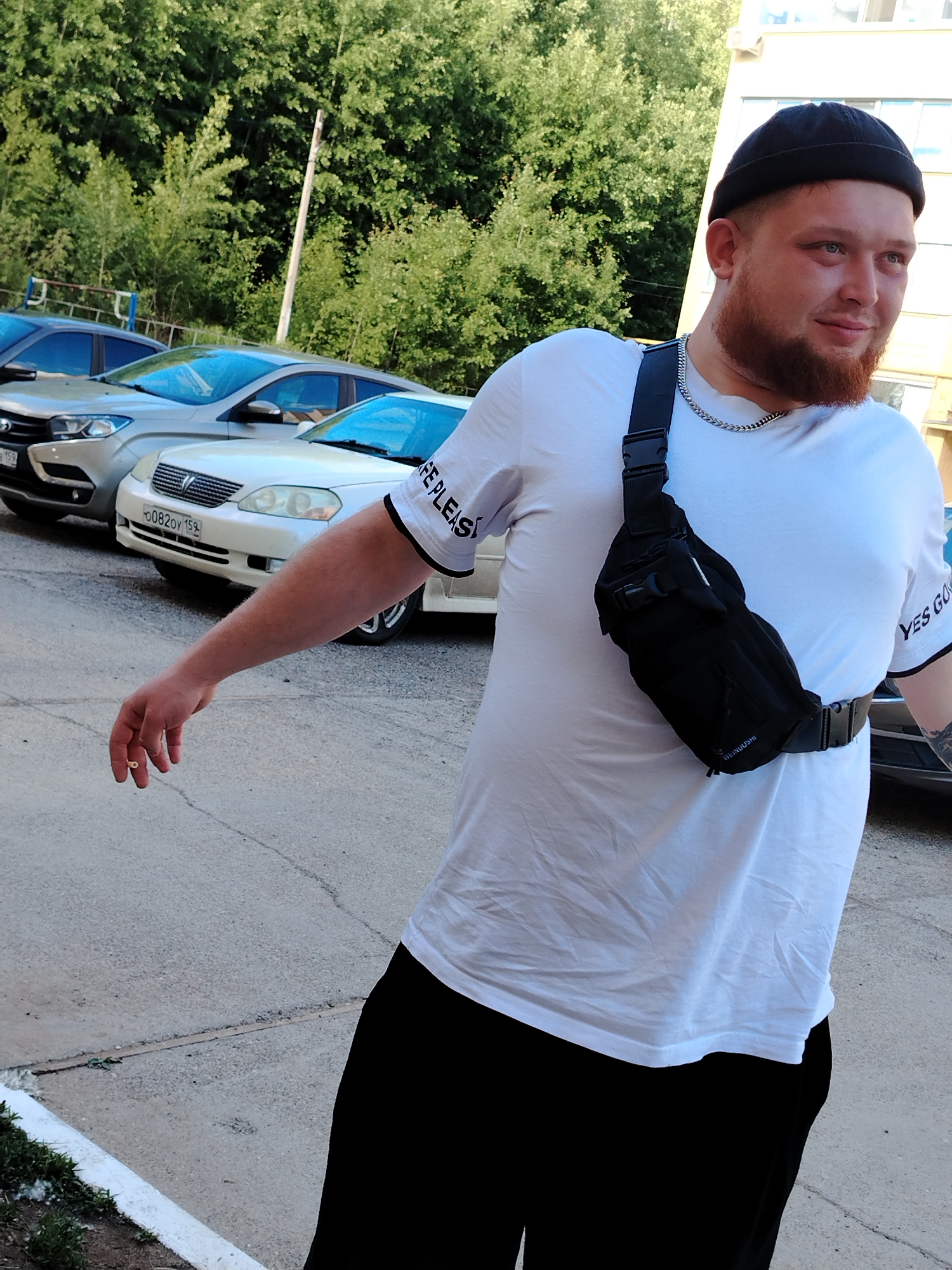 A man with a red beard and wearing a black beanie, white t-shirt with black trim, black pants, and a black fanny pack is standing outdoors. He is looking to the right with a slight smile, and his left arm is extended to the side. In the background, there are three cars parked on a paved surface, and green trees are visible in the distance. The license plate on the white car in the middle reads "O082OY 159". The t-shirt has words printed on the sleeves. The photograph was taken in the city of Noviy, Russia.